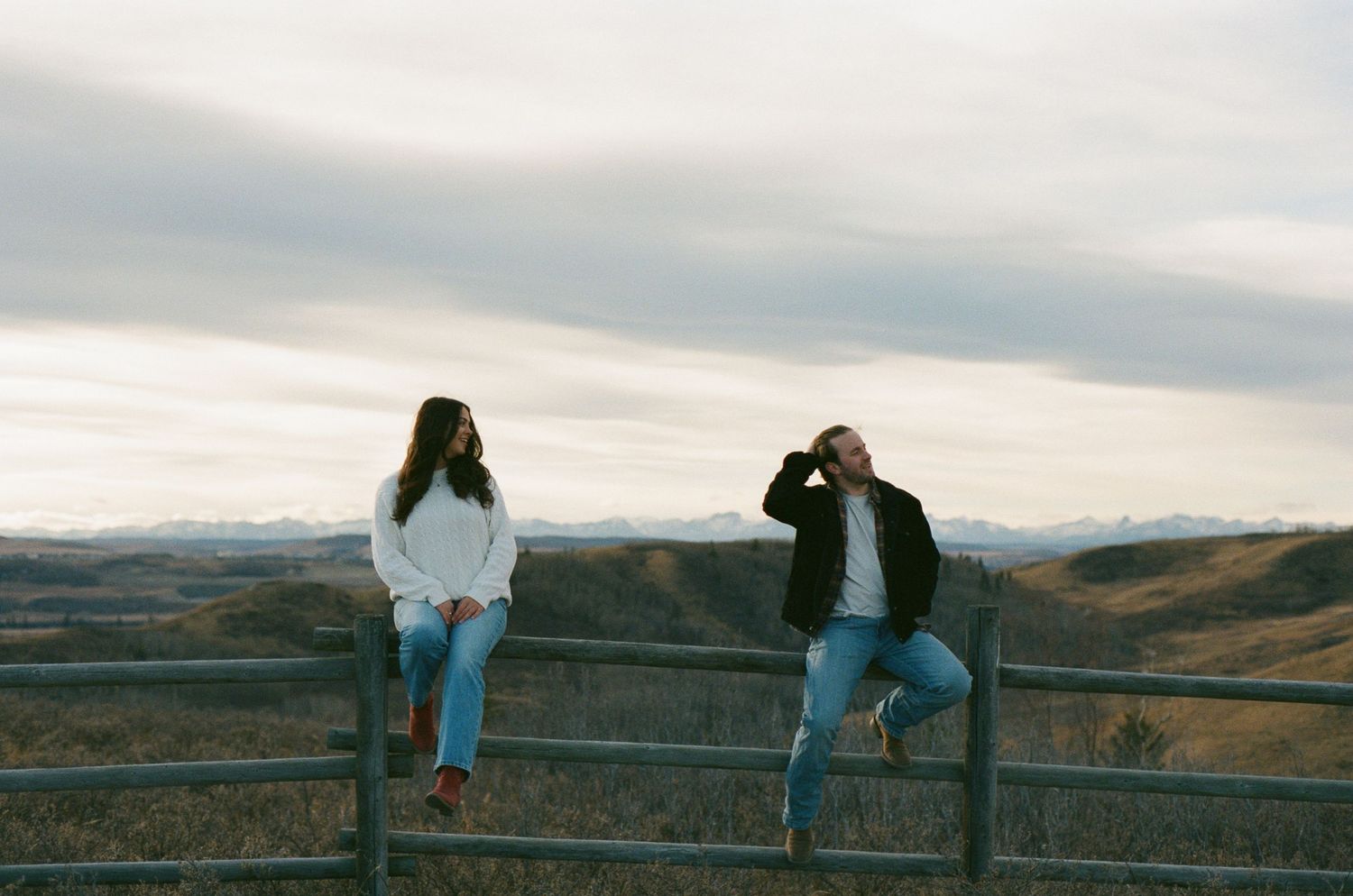 Two people sit on a wooden fence overlooking a scenic mountain landscape at sunset.