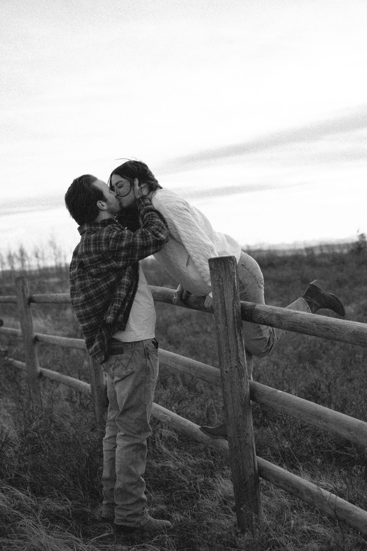 A romantic couple shares a tender moment leaning against a rustic wooden fence in black and white.