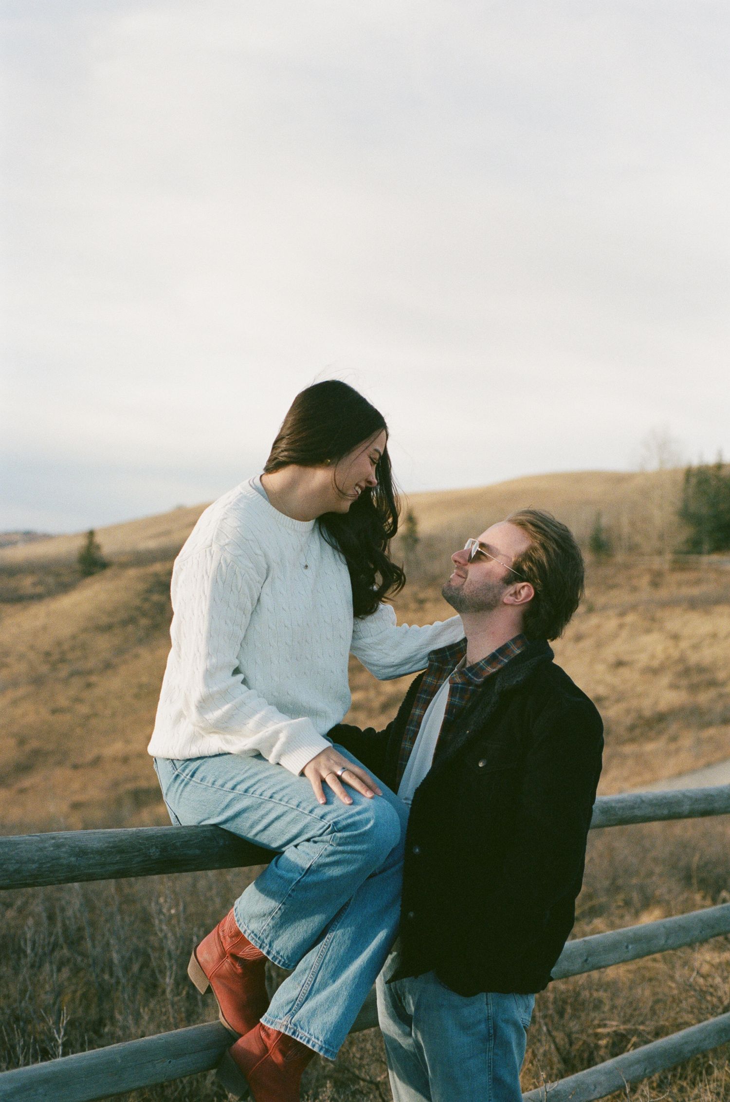 A couple shares a romantic moment while sitting on a wooden fence in a rural setting.