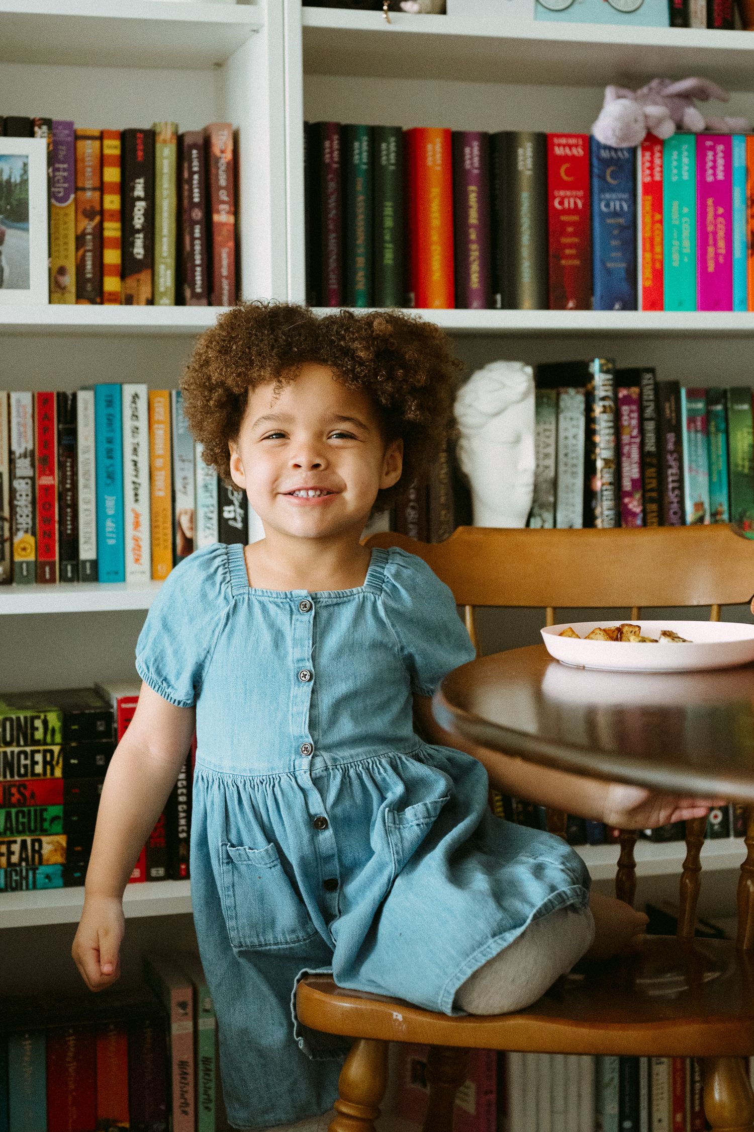 A smiling young girl in a denim dress sits on a wooden chair in front of a colorful bookshelf.