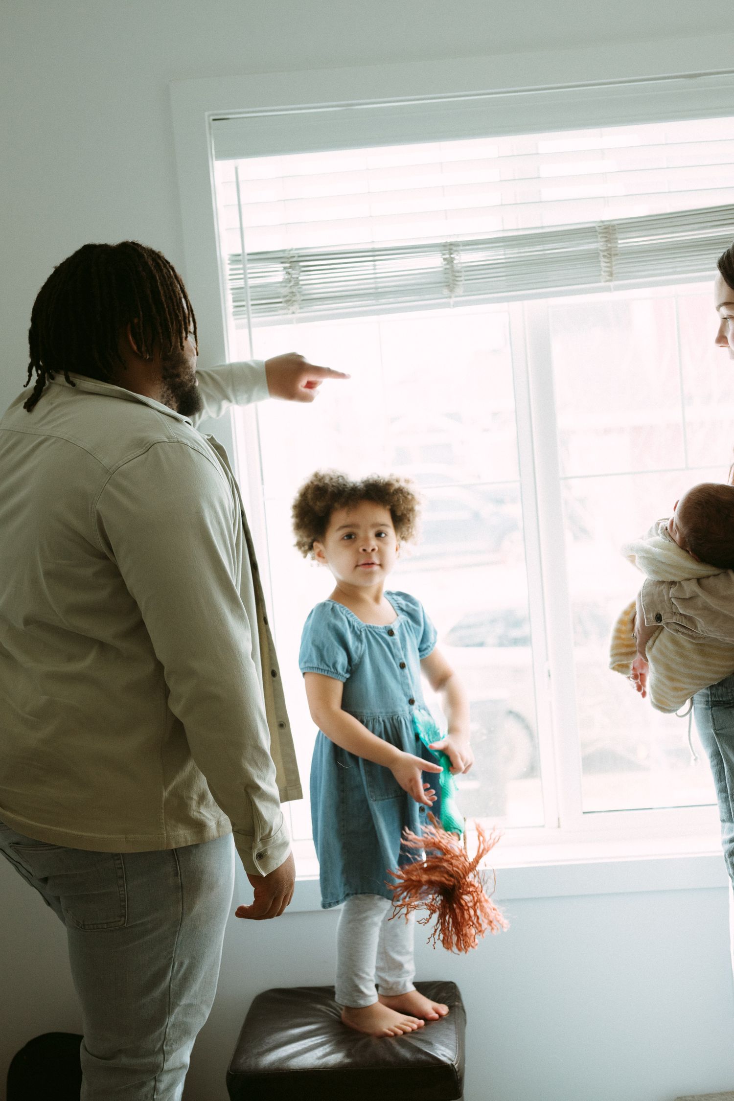 A father points out a window to his young daughter standing on a ottoman, while mother holds baby nearby.