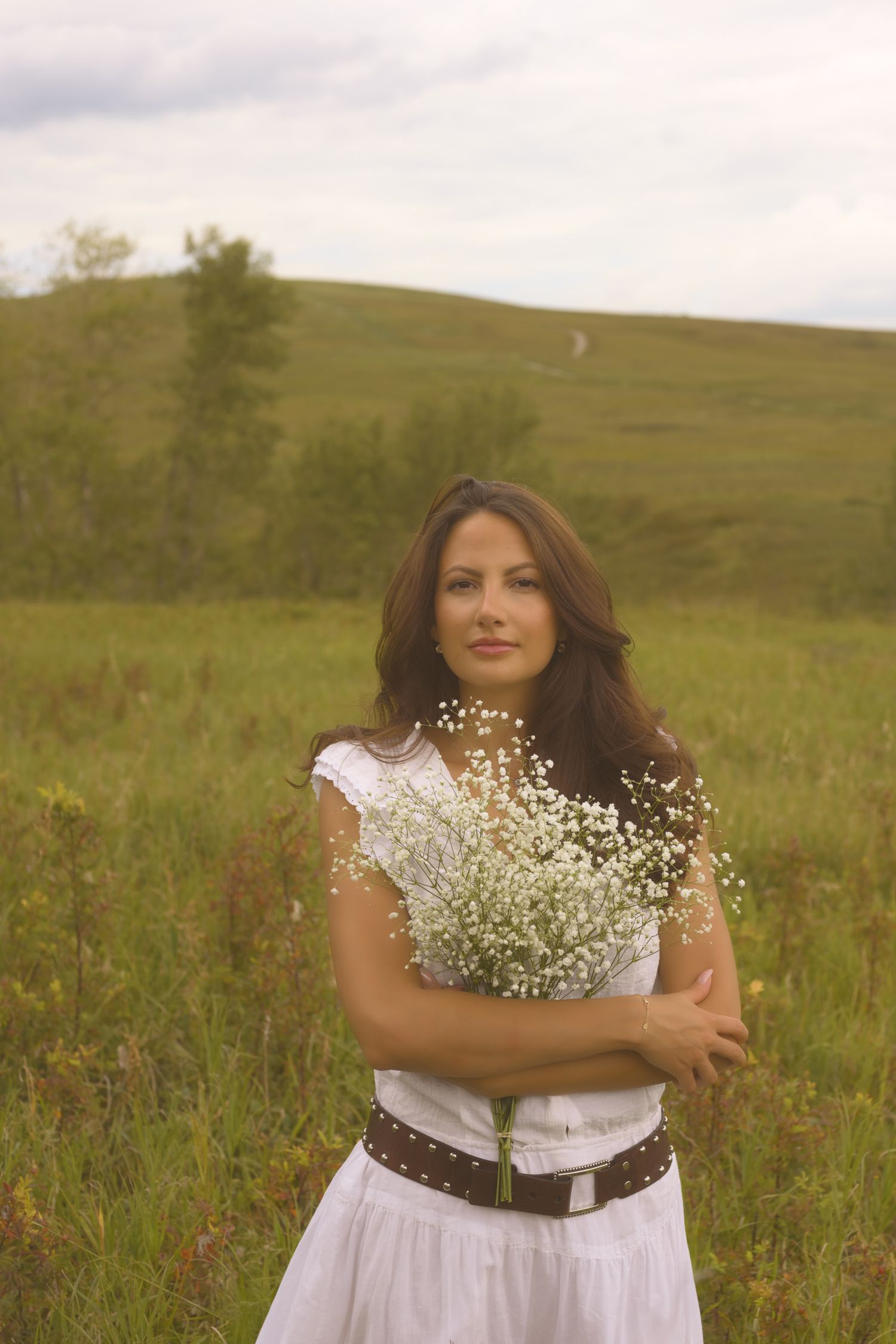 A figure in white holds baby's breath flowers against rolling hills and cloudy skies.