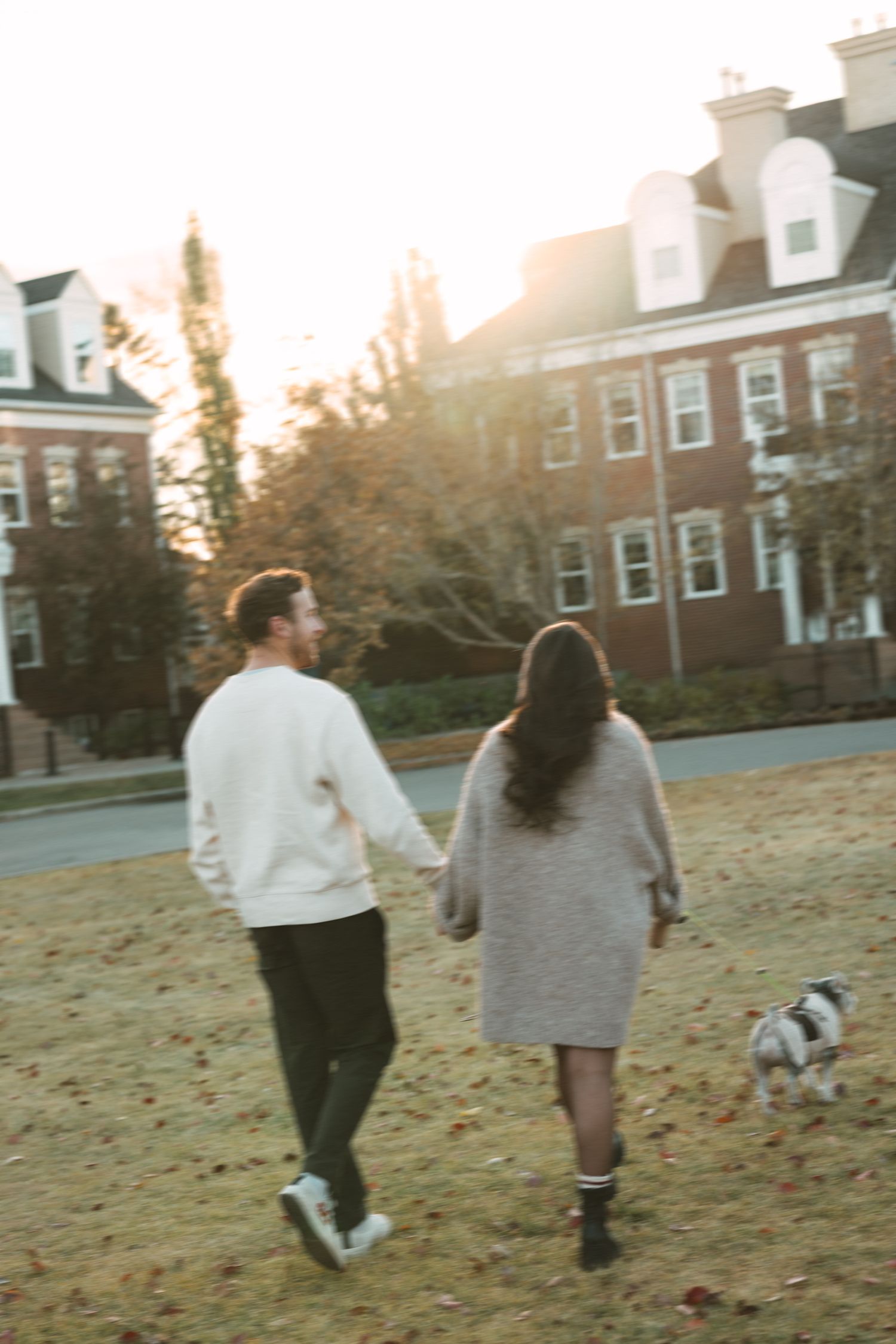 Two people walk with a small dog on a grassy lawn at sunset with brick buildings in the background.