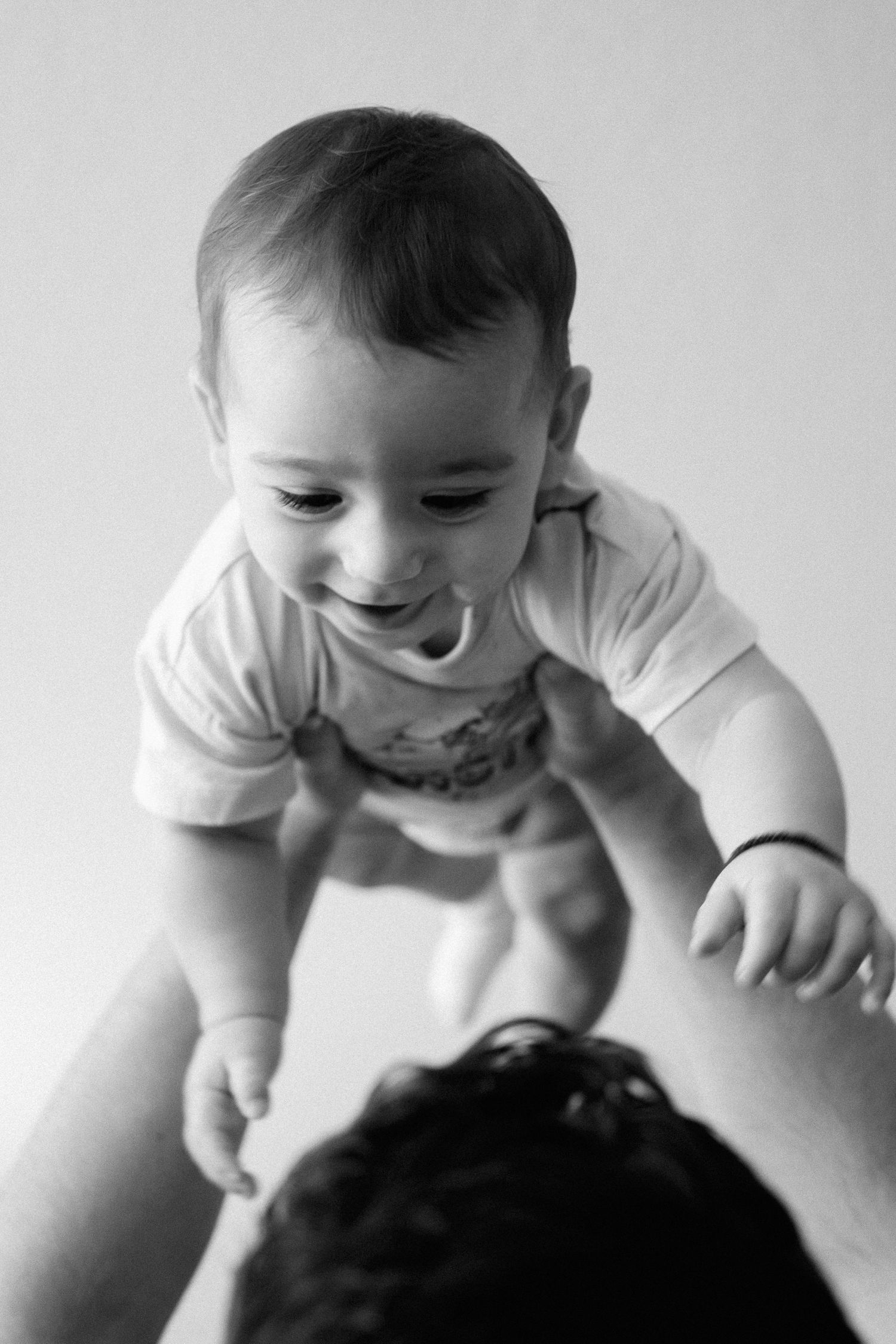 A baby in white clothing smiles with delight in black and white photography.