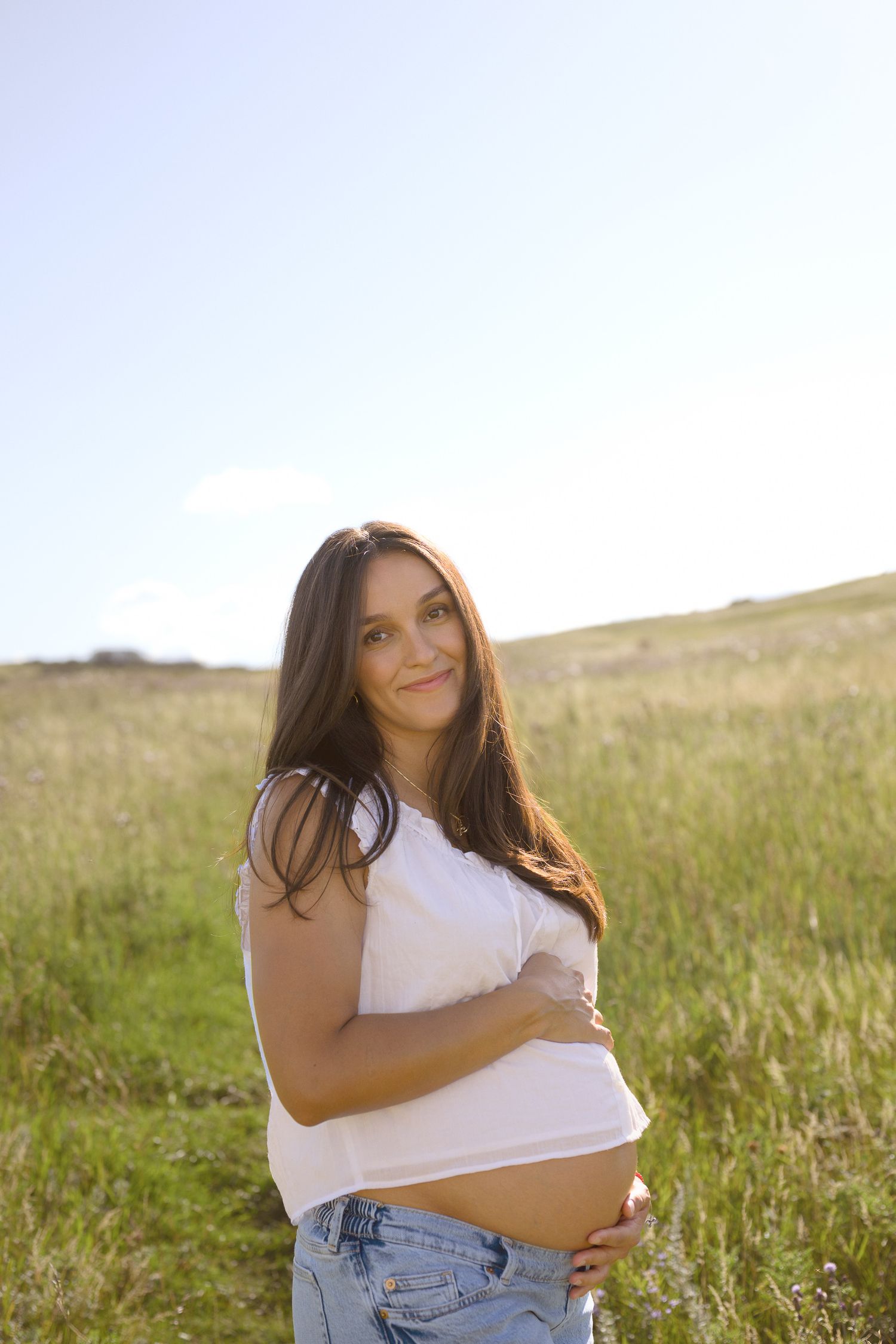 Woman in white top and jeans poses joyfully in a sunlit meadow.