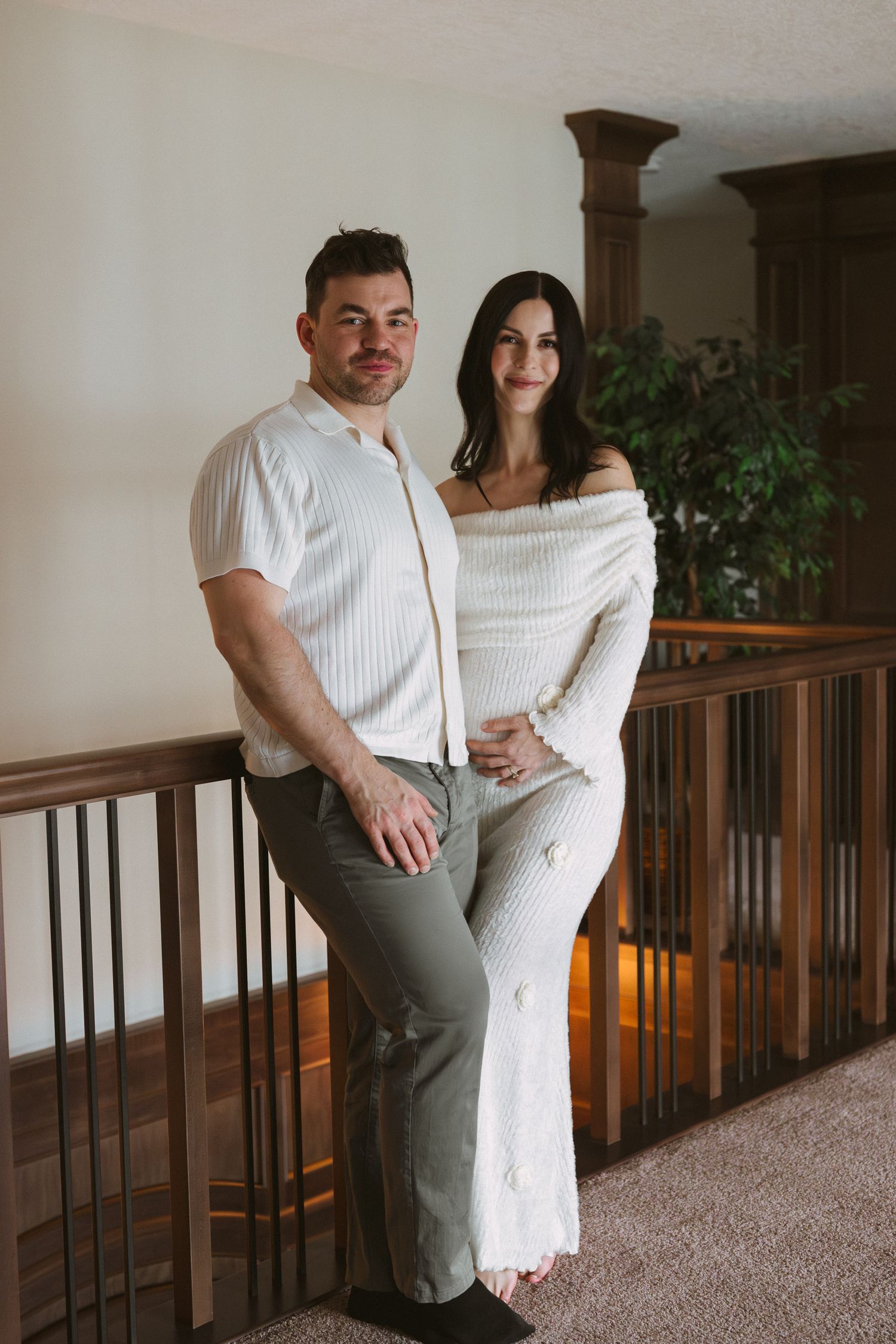 A couple poses together on an indoor balcony, both wearing white outfits, with wooden railings and greenery in the background.