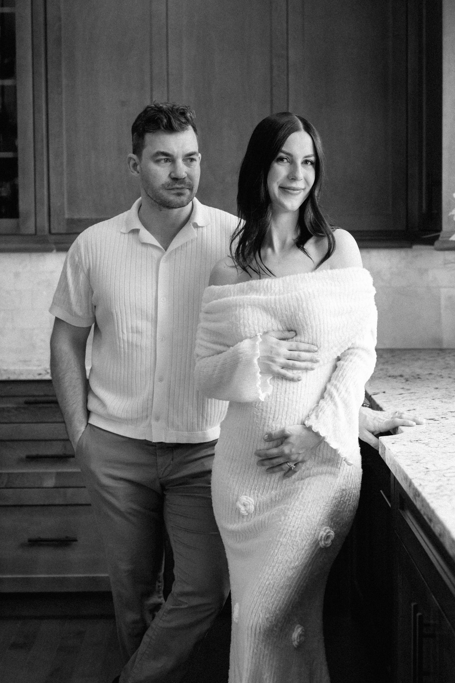 Black and white maternity photo of a couple posing in a kitchen, woman in off-shoulder dress cradling baby bump.