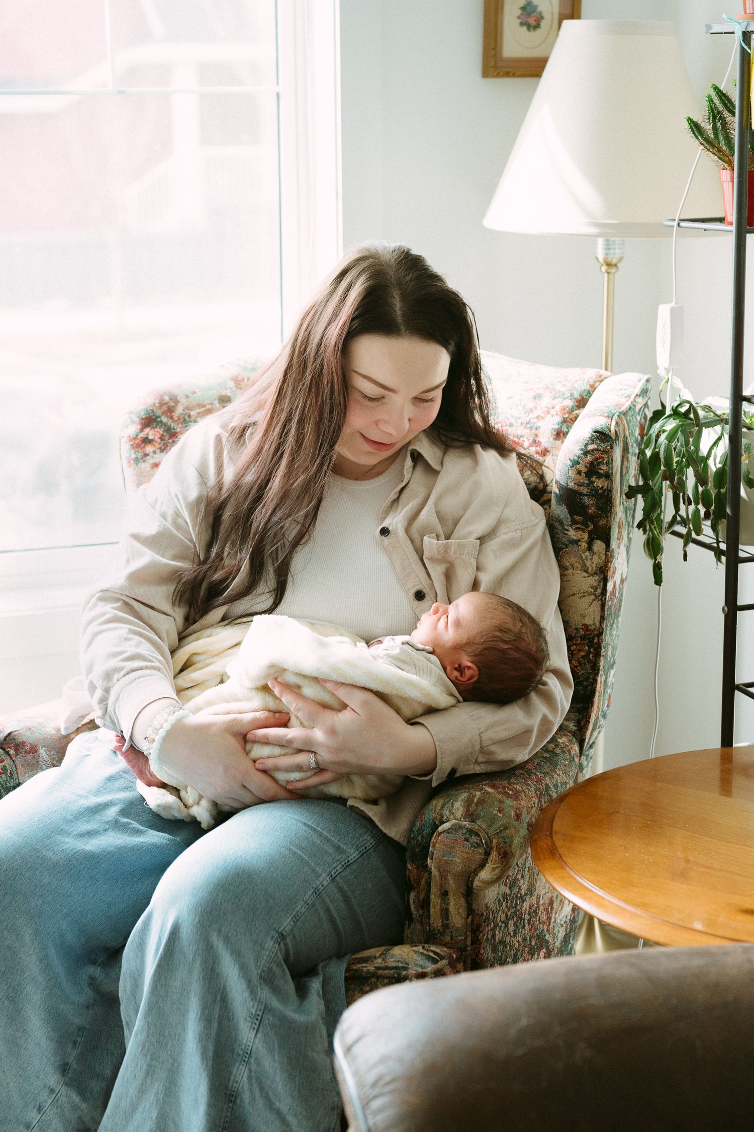 A mother with long dark hair sits in a floral armchair, tenderly cradling her newborn baby wrapped in a white blanket.