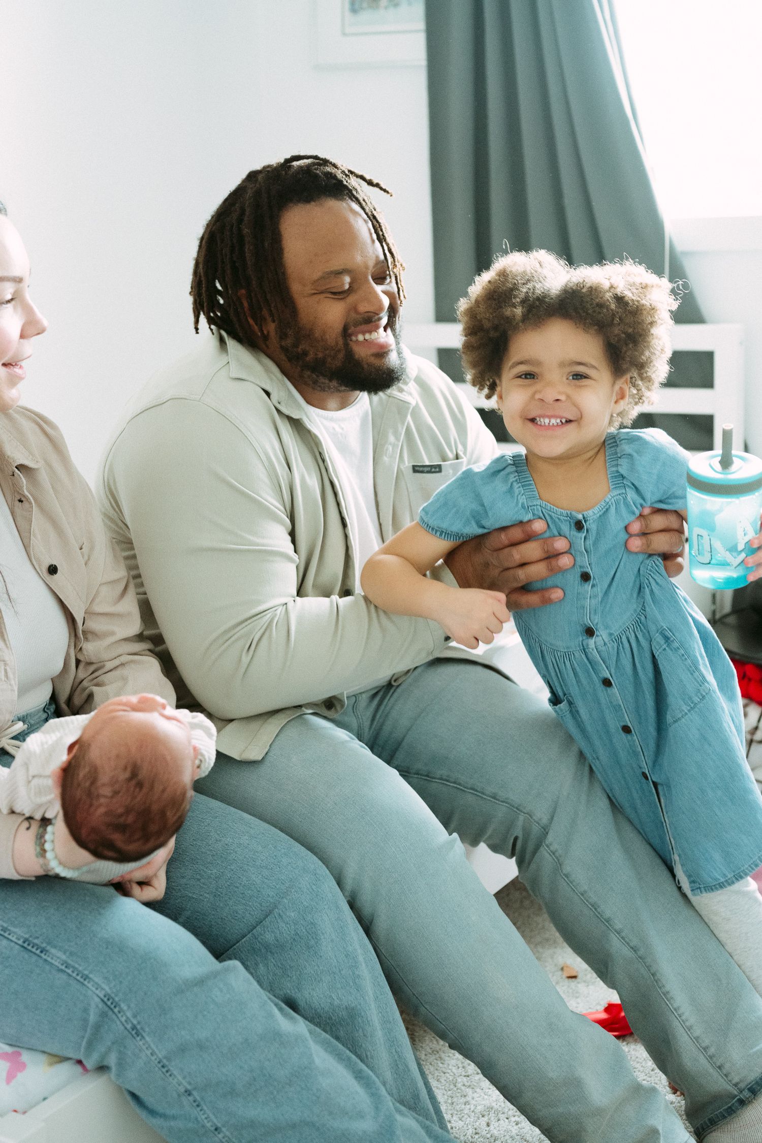 A joyful family sits together, with a dad playing with his curly-haired daughter in a blue dress while mom holds a newborn.