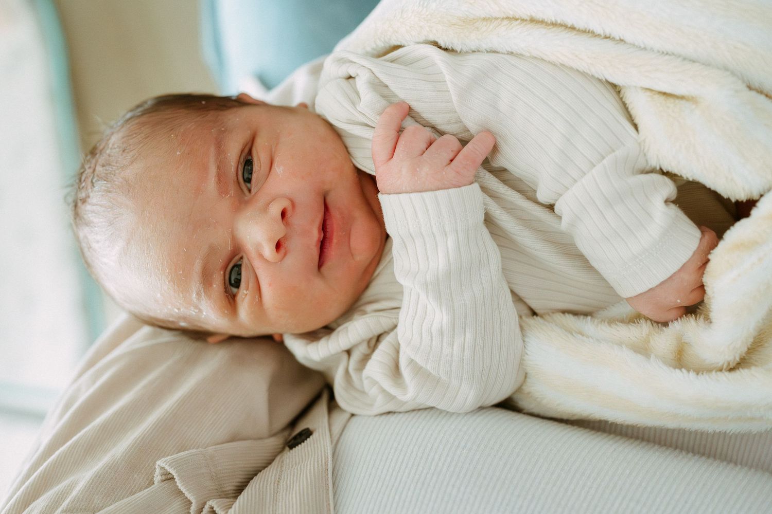 Adorable newborn baby in white outfit looking up while being held, wrapped in soft blanket.