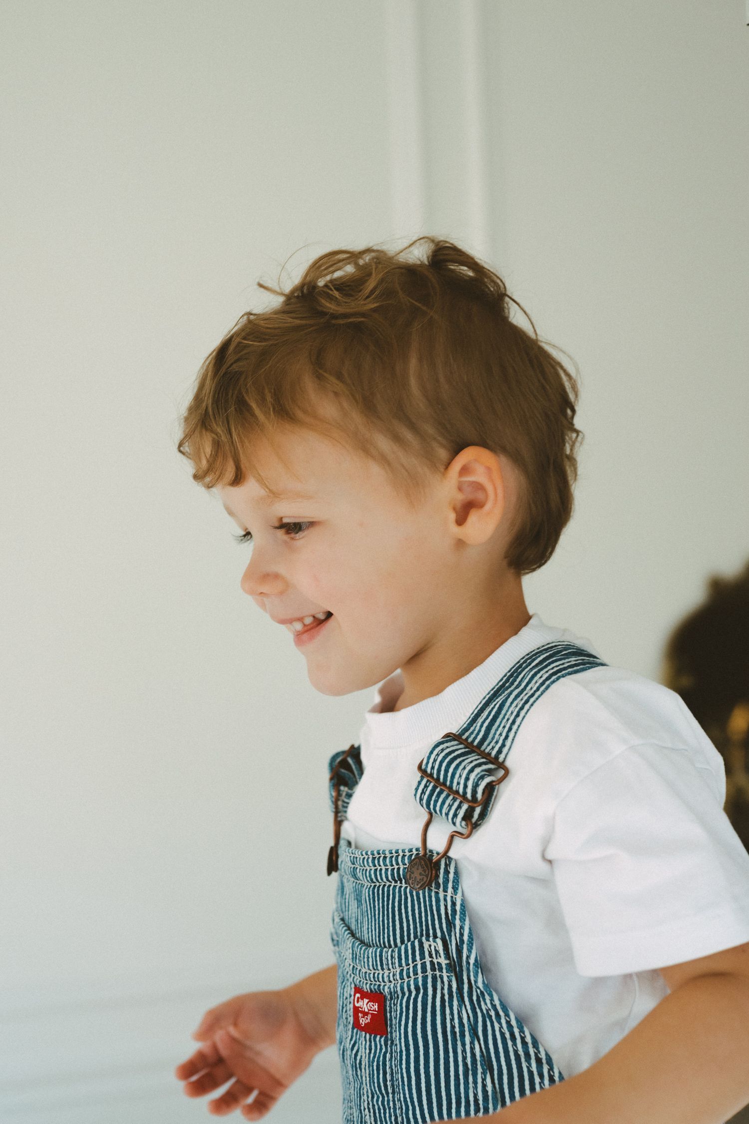 A young child in striped overalls smiles playfully while being held against a white wall background.