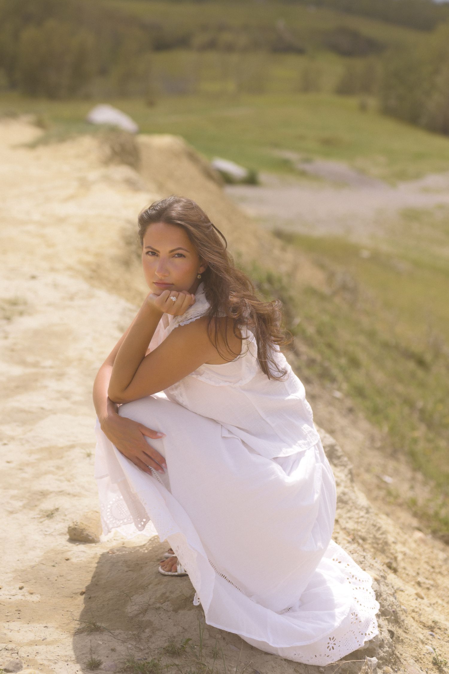 Ethereal portrait in white dress sitting on rocky terrain with sunlit hills in background.