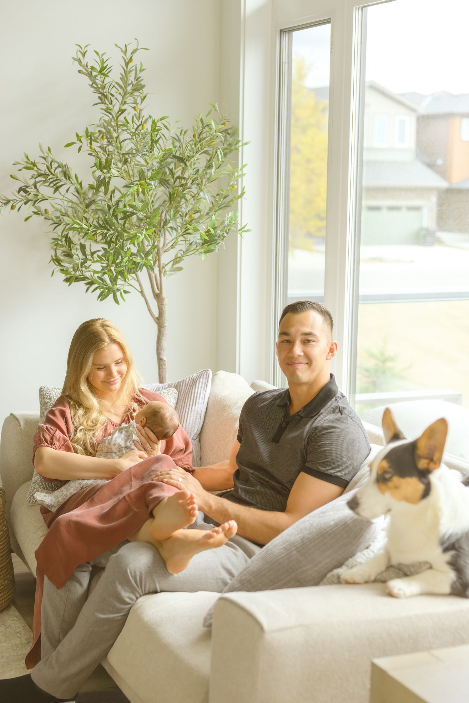 Family sits on white couch with their dog near a potted indoor tree.