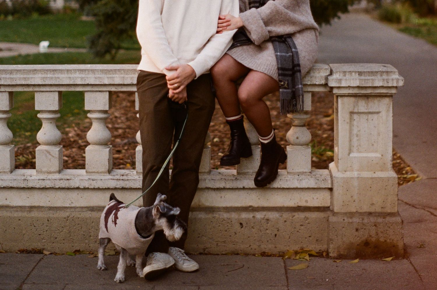 A black and white dog on a leash sits near someone sitting on a stone balustrade on an autumn day.