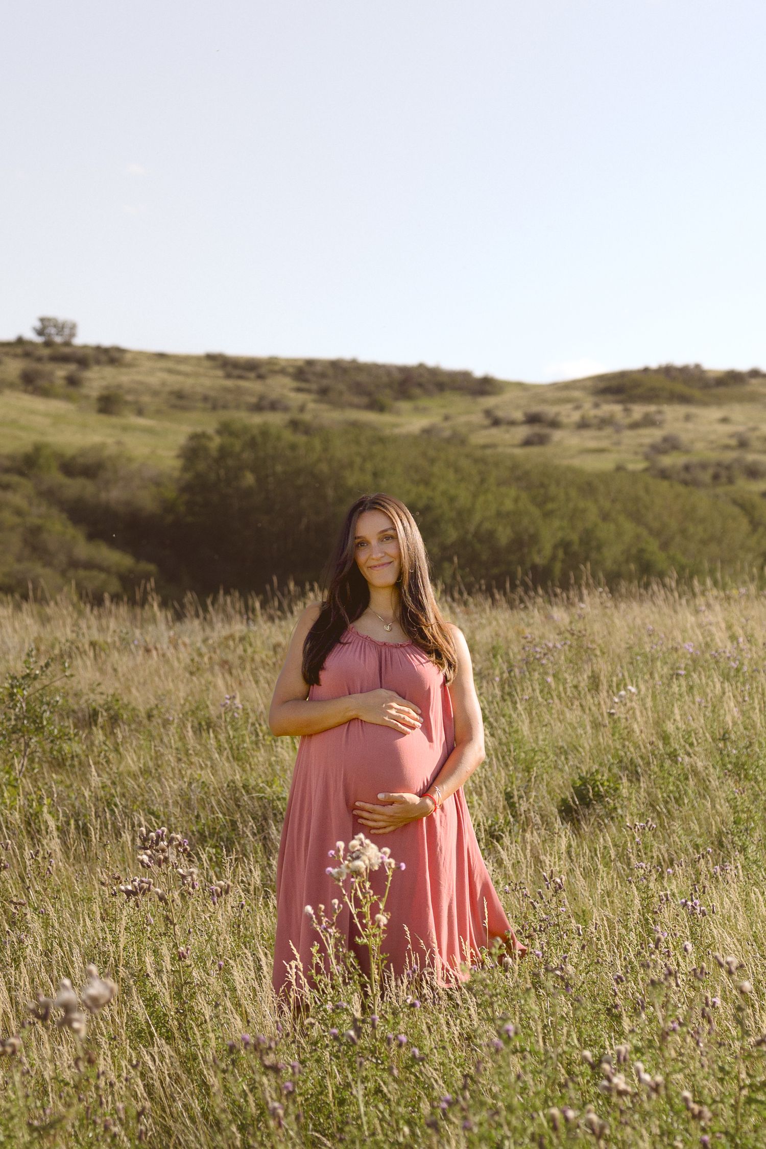 A pregnant woman in a pink dress stands in a sunlit meadow with rolling hills.