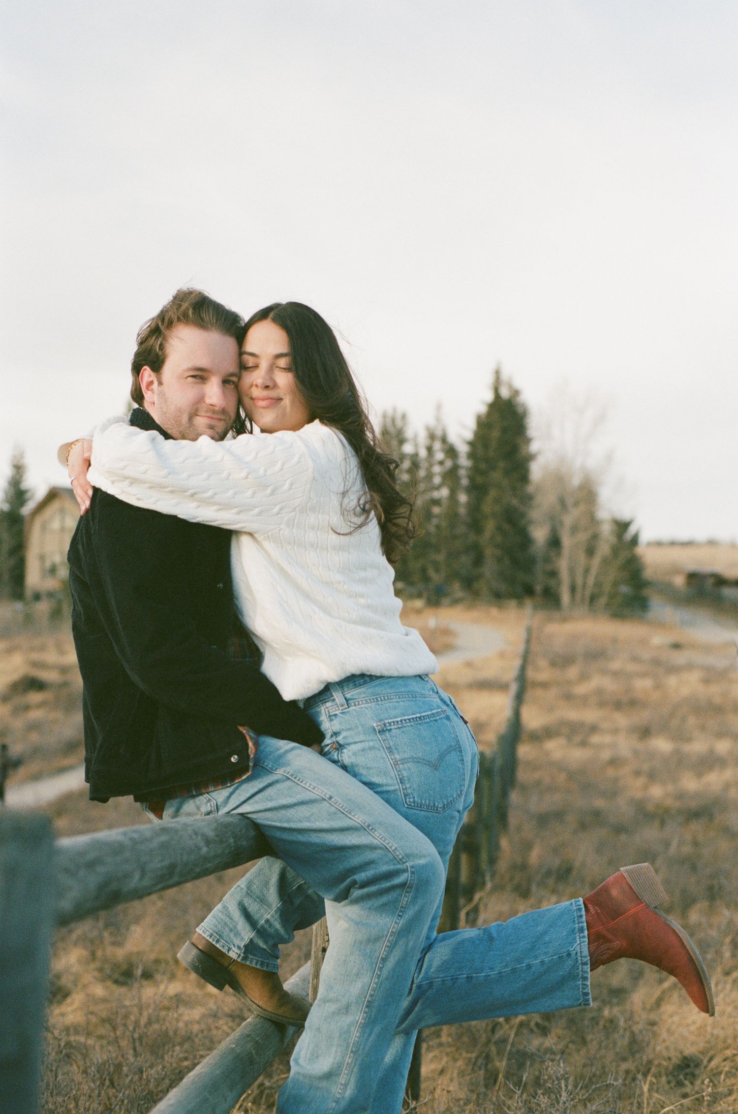 A playful couple embraces while sitting on a wooden ranch fence in a rural autumn landscape.