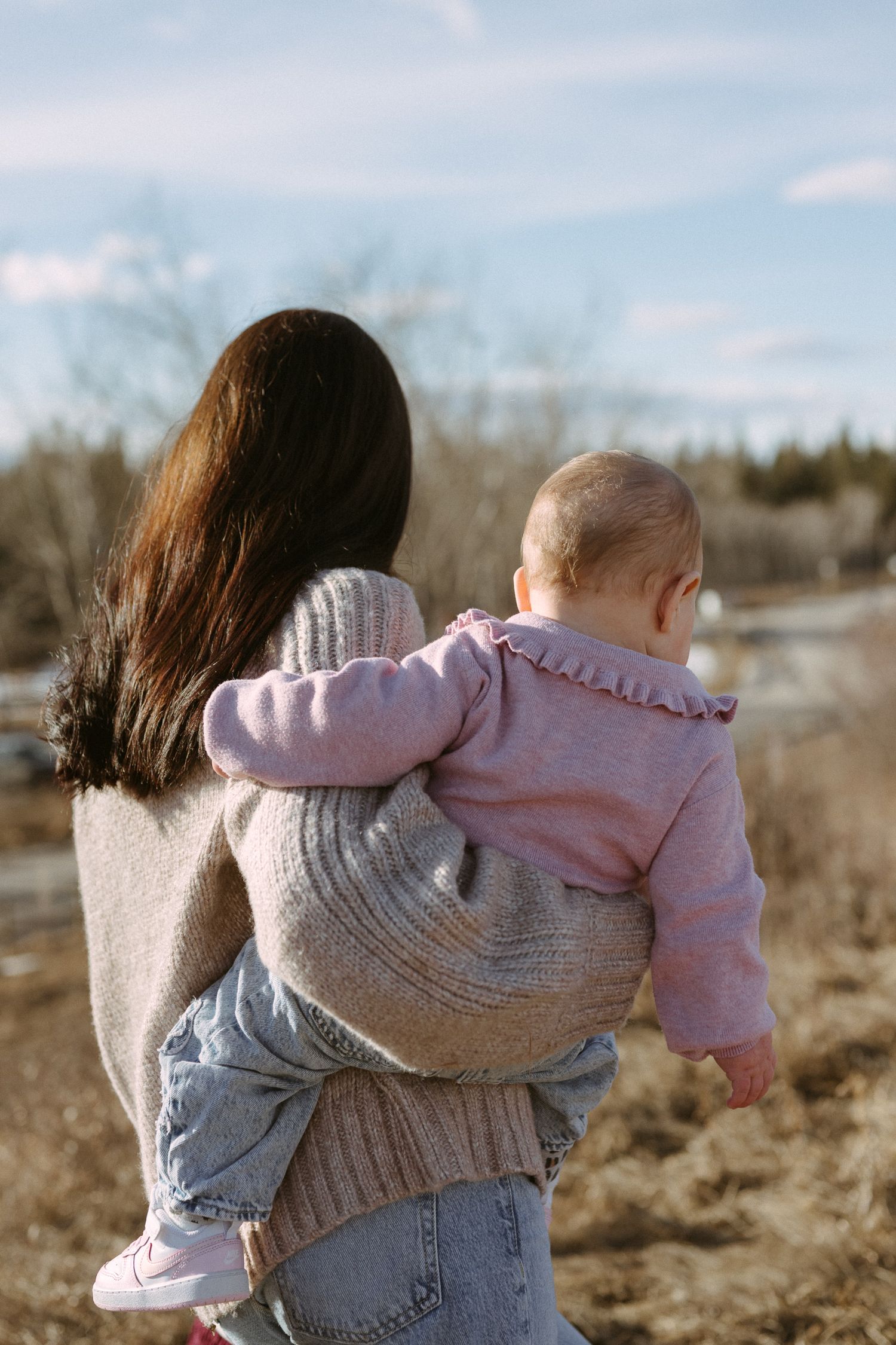 A person in a knit sweater holds a baby wearing pink while standing in a sunny outdoor setting.