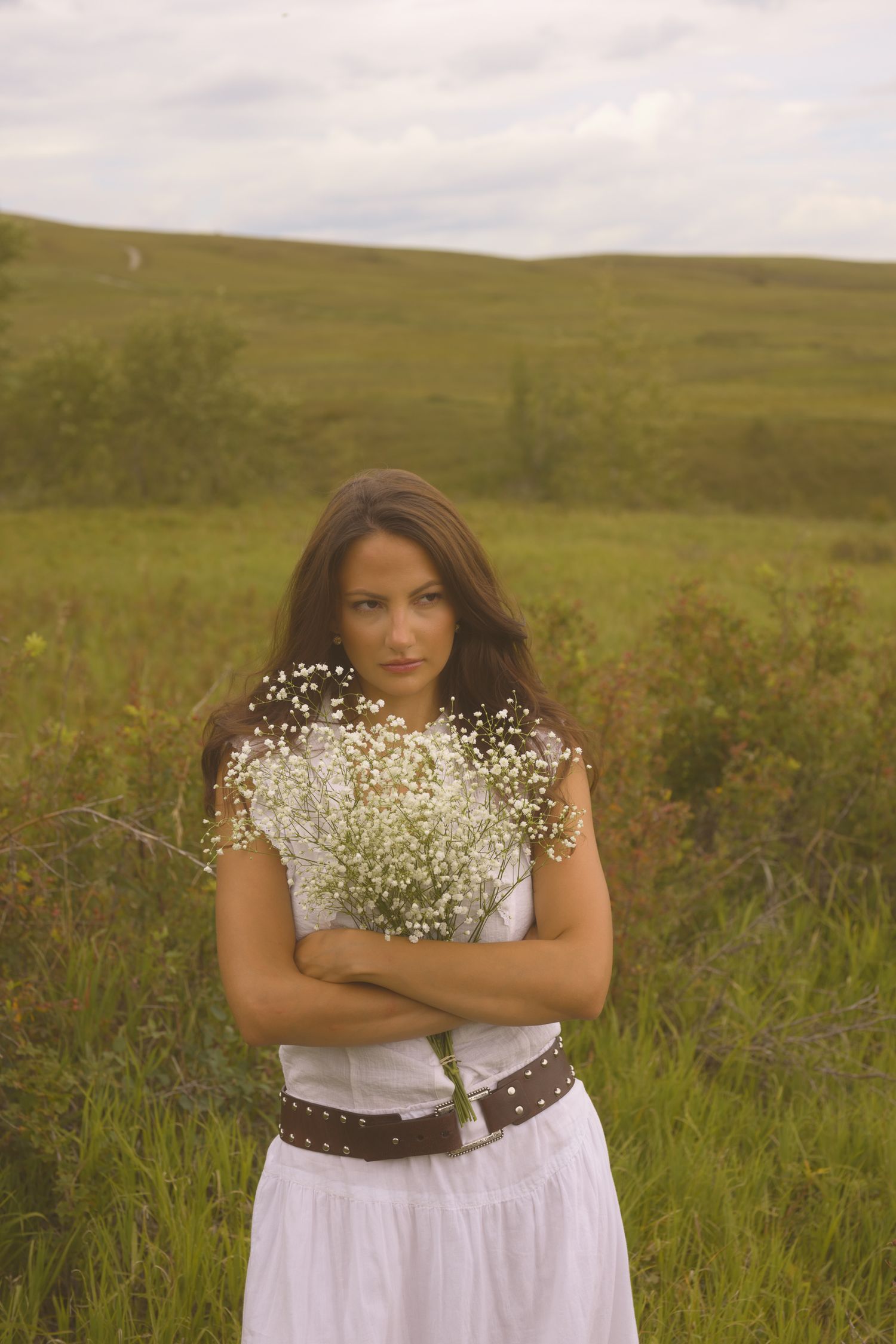 Someone wearing a white dress and brown belt holds baby's breath flowers in a grassy field.