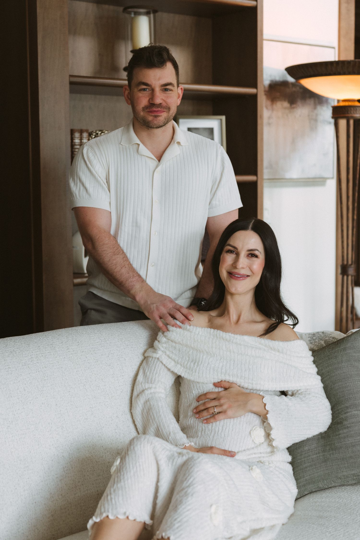 Happy couple in white outfits posing on cream sofa, pregnant woman seated with partner standing behind her.