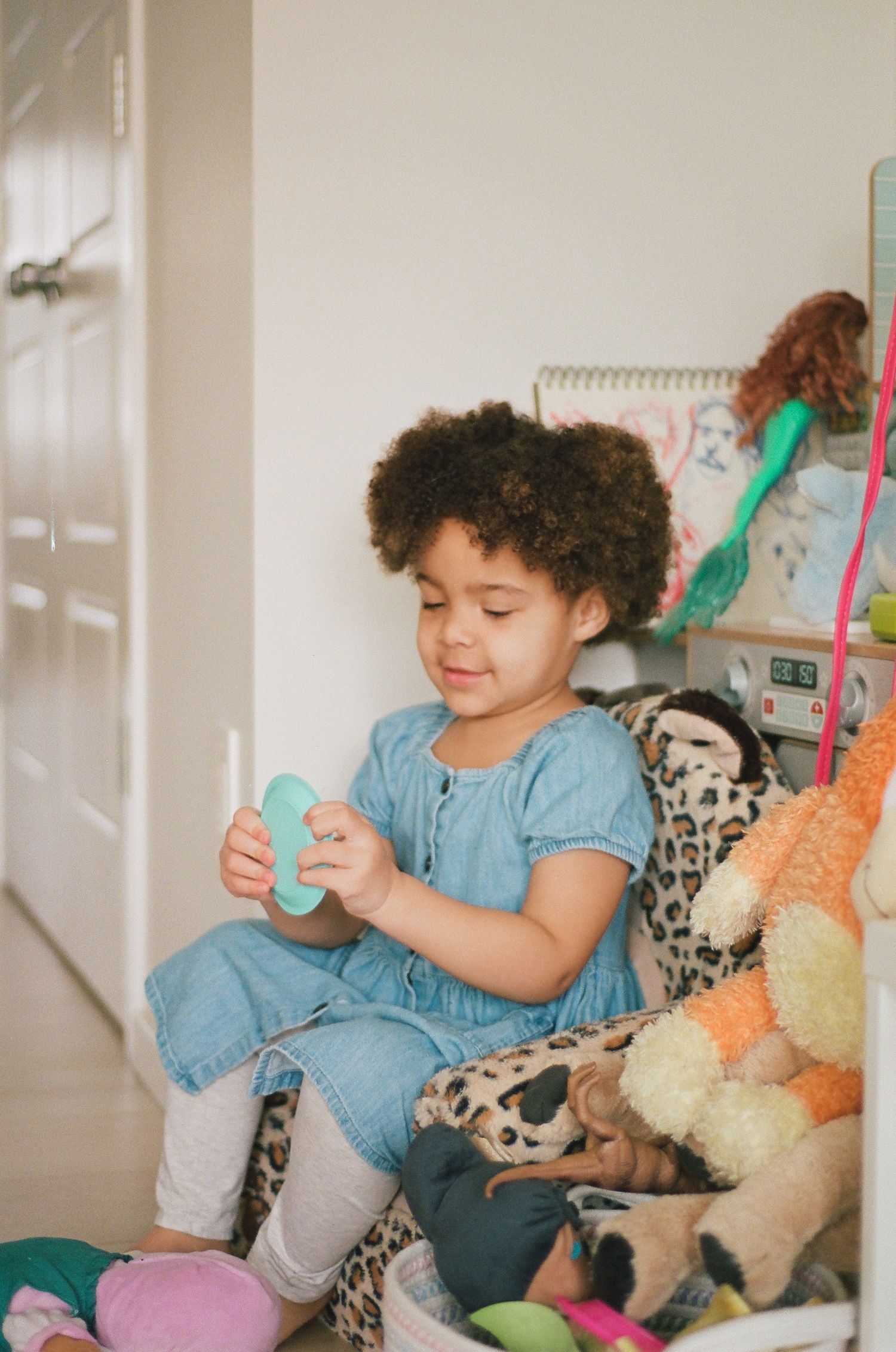 Young girl with curly hair in blue dress playing with toys while sitting in a leopard print chair.