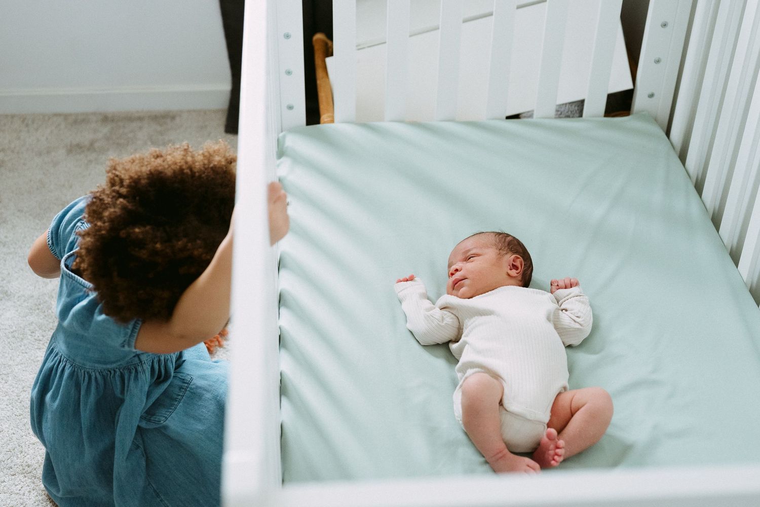 A curly-haired toddler peers over a white crib rail at a sleeping newborn baby.