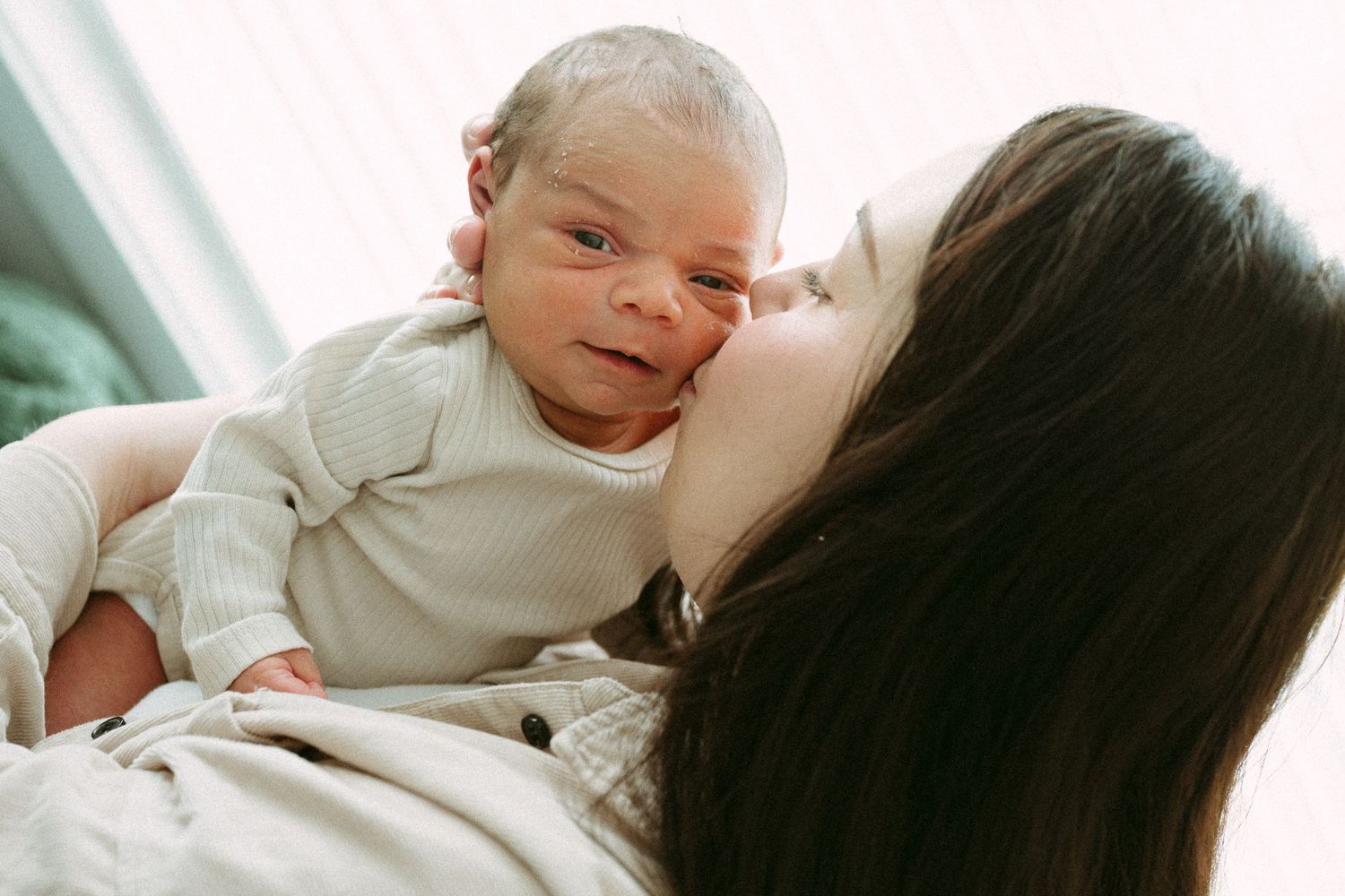 A mother tenderly kisses her newborn baby, who looks up curiously in a soft, bright room.