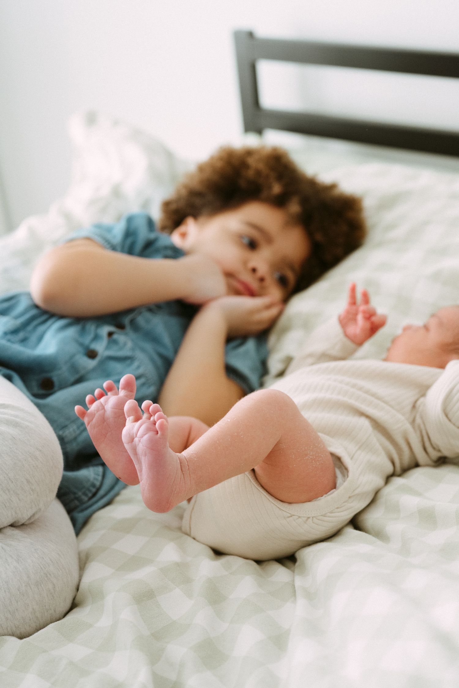 Close-up of a curly-haired child lying on a bed smiling at a newborn baby with tiny feet in the foreground.
