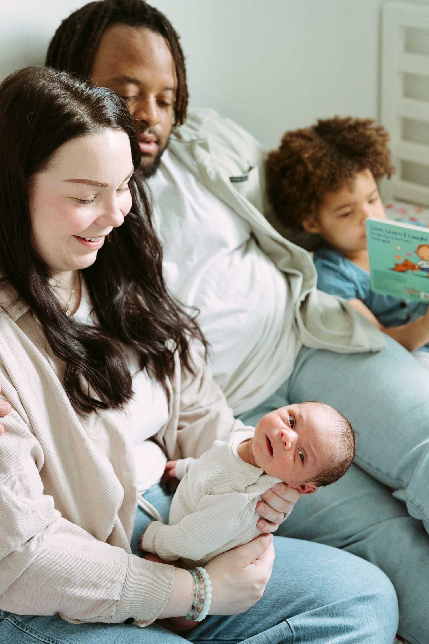 A multiracial family sits together, with a smiling mother holding a newborn baby while father and toddler read a book.