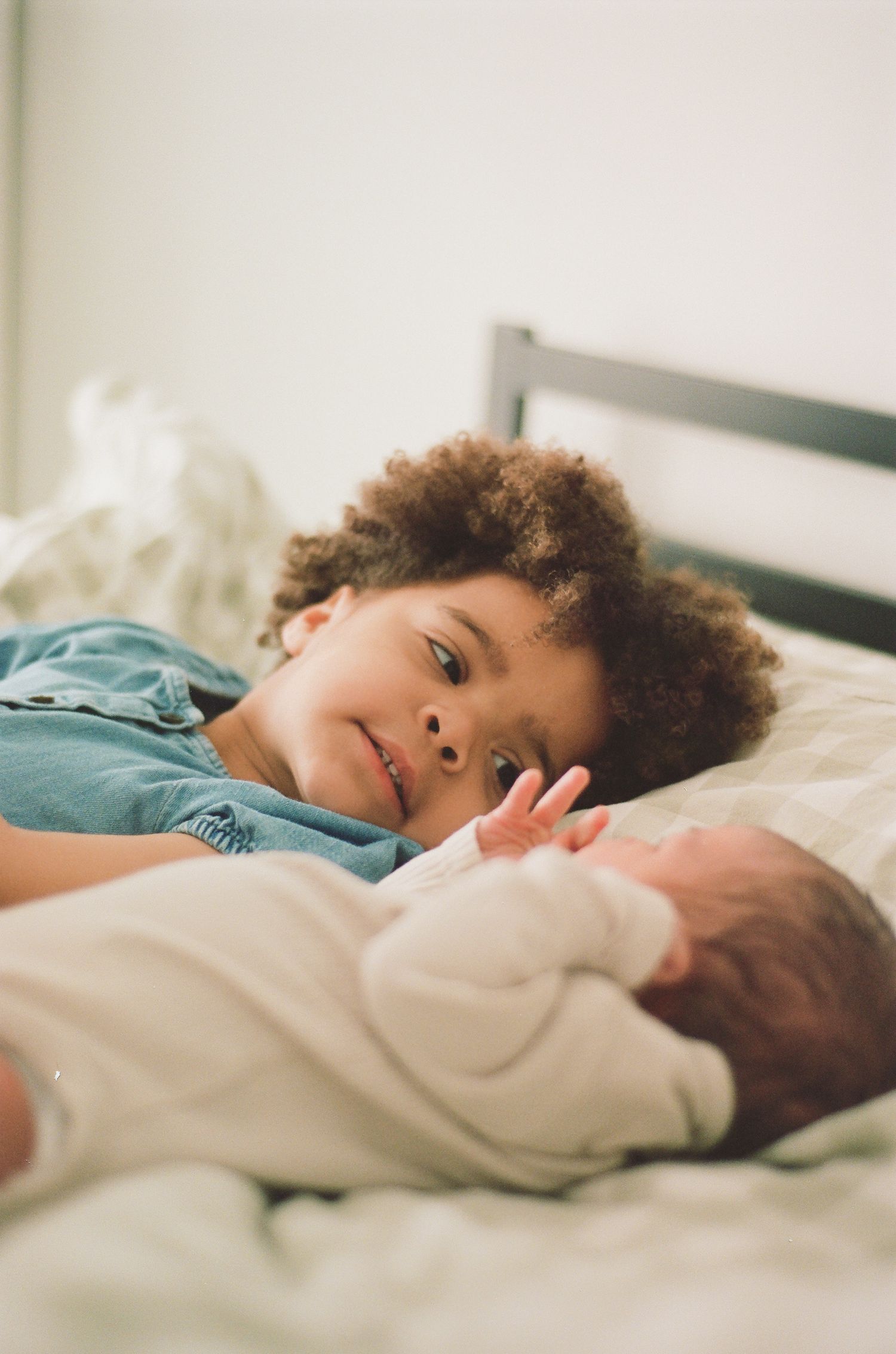 A curly-haired toddler smiles warmly while lying next to a newborn baby on a white bed.