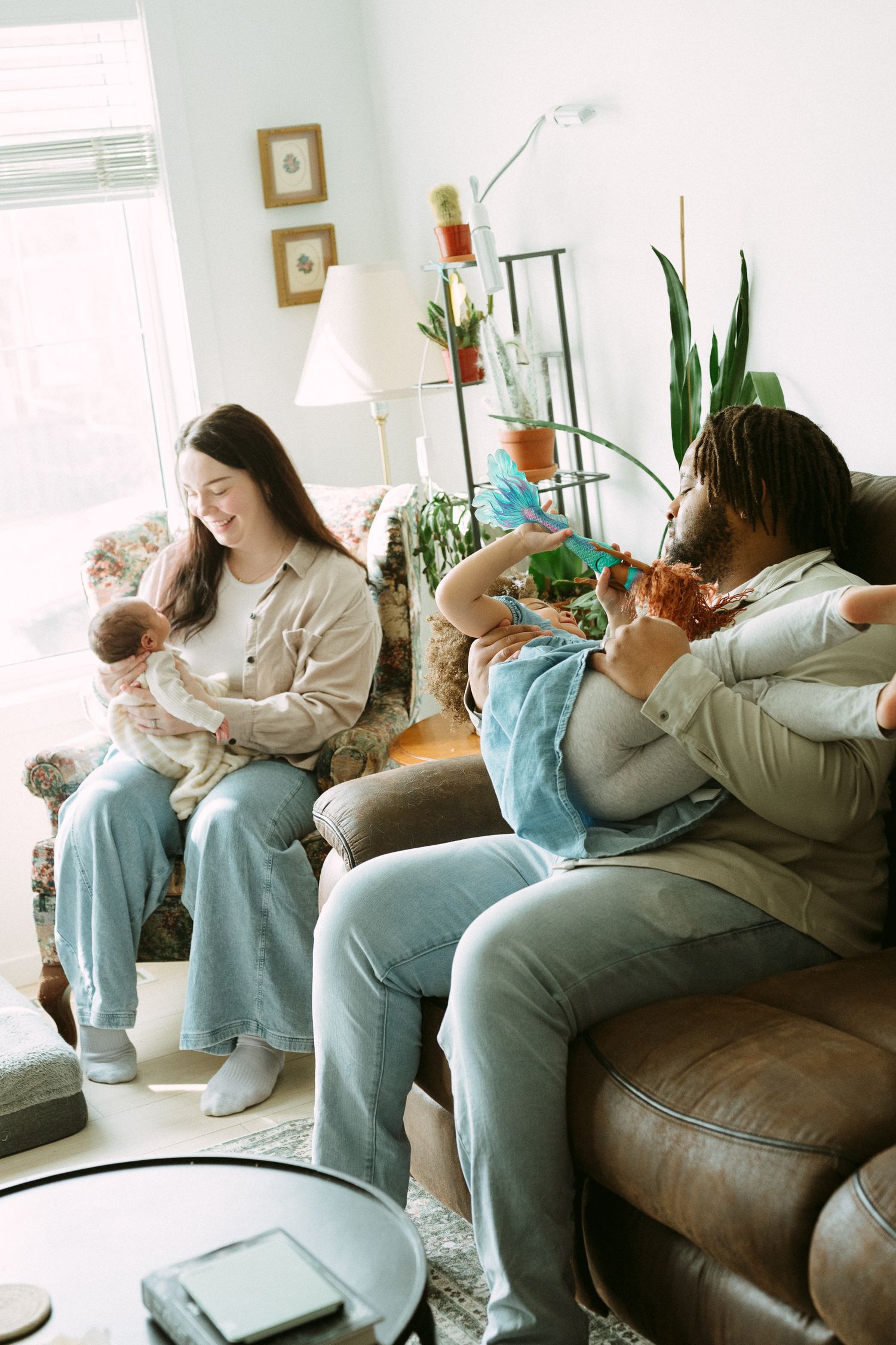 Interracial family relaxing on couch at home, mother holding newborn baby while father plays with toddler daughter.