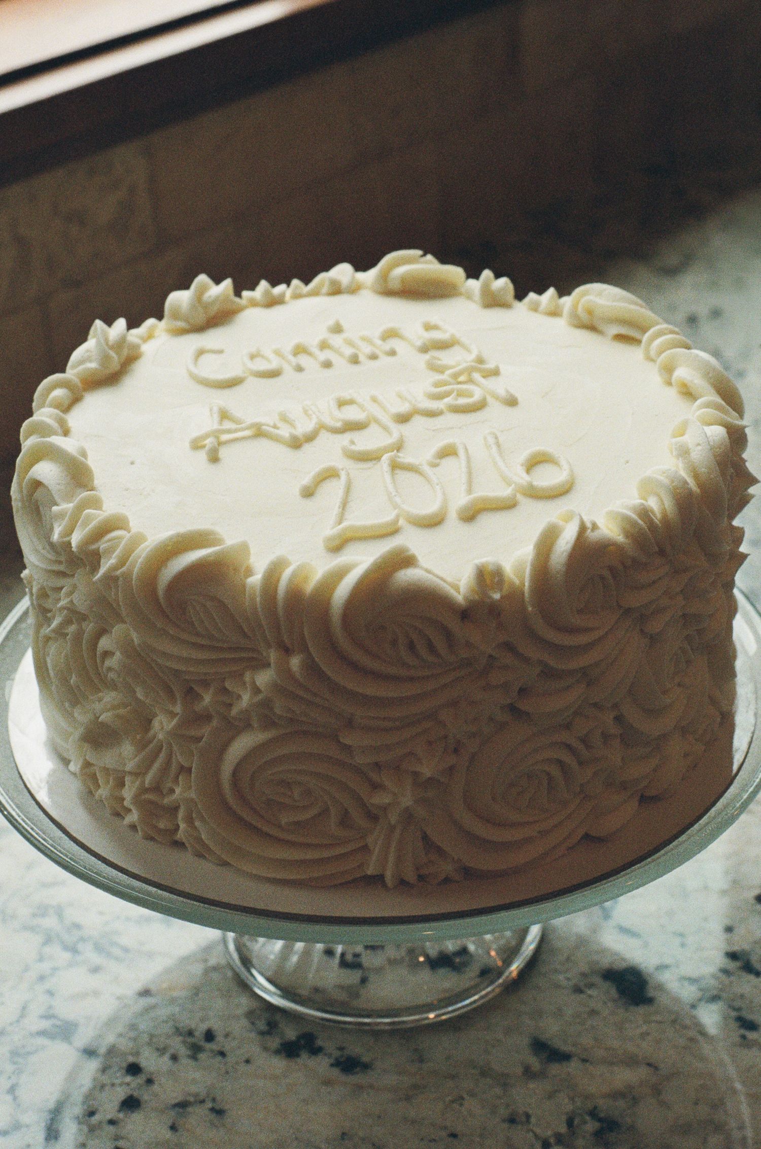 White frosted celebration cake on glass stand reading Coming August 2024 with rosette piping decoration.
