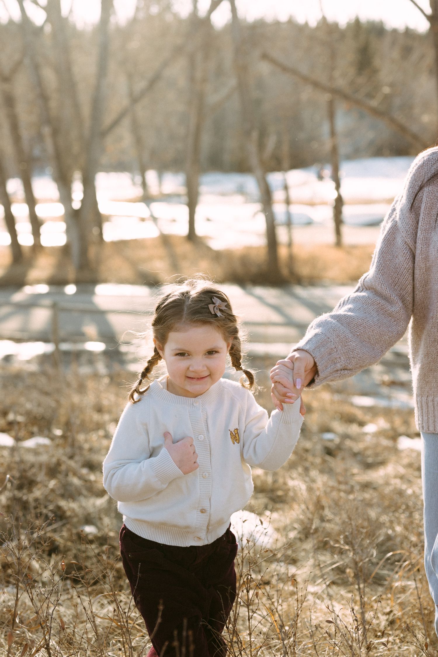 A small child in a white sweater walks through a snowy winter landscape holding an adult's hand.