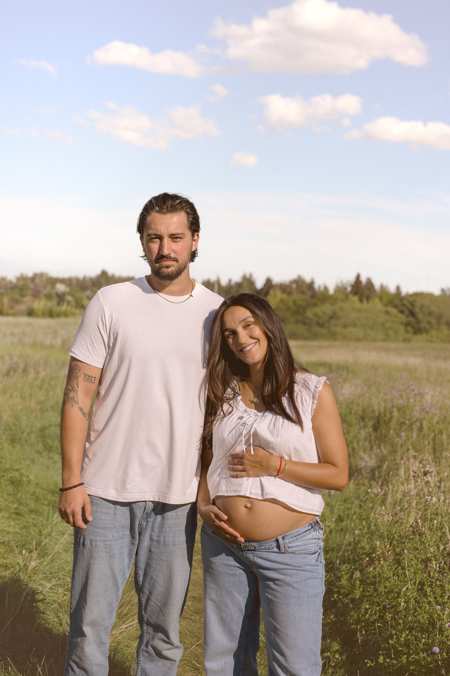 Couple in casual denim and white outfits standing together in a sunny meadow.