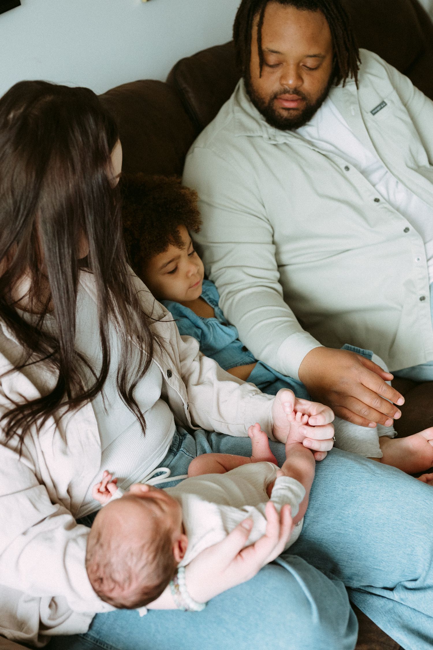A loving family sits together on a couch, with parents and toddler admiring their newborn baby.