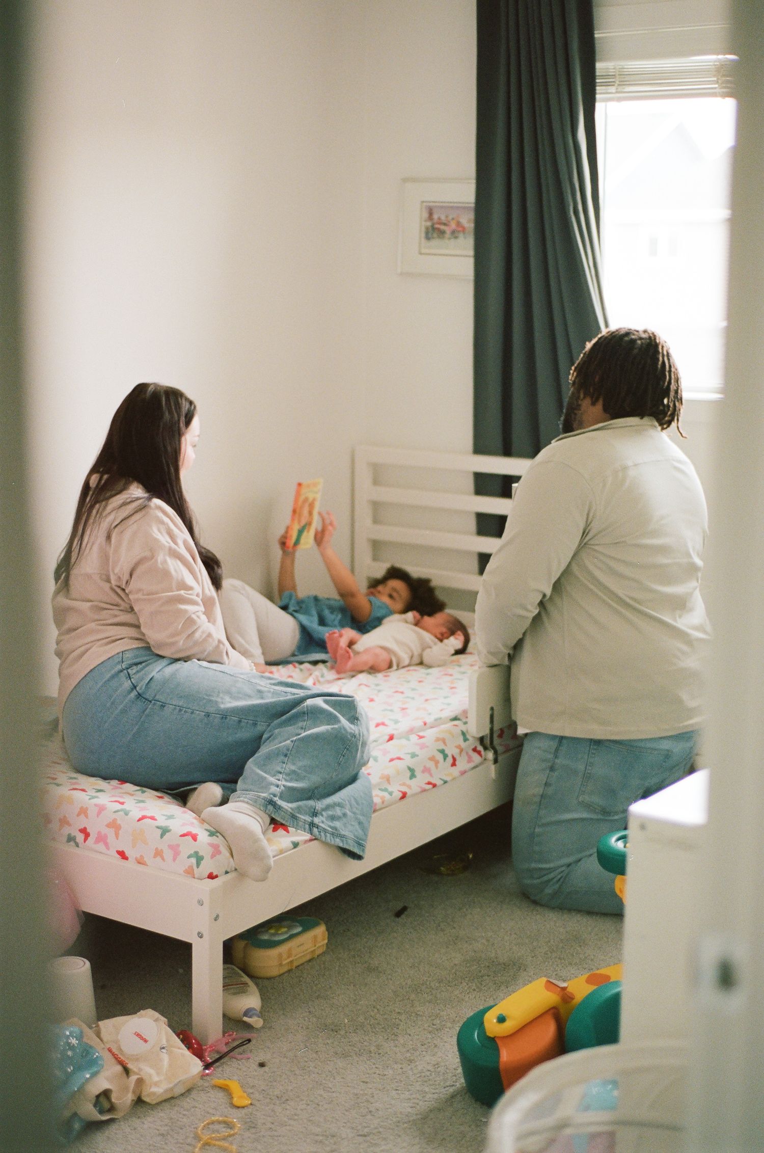 Two adults sit beside a child lying on a white toddler bed in a cozy bedroom with teal curtains.