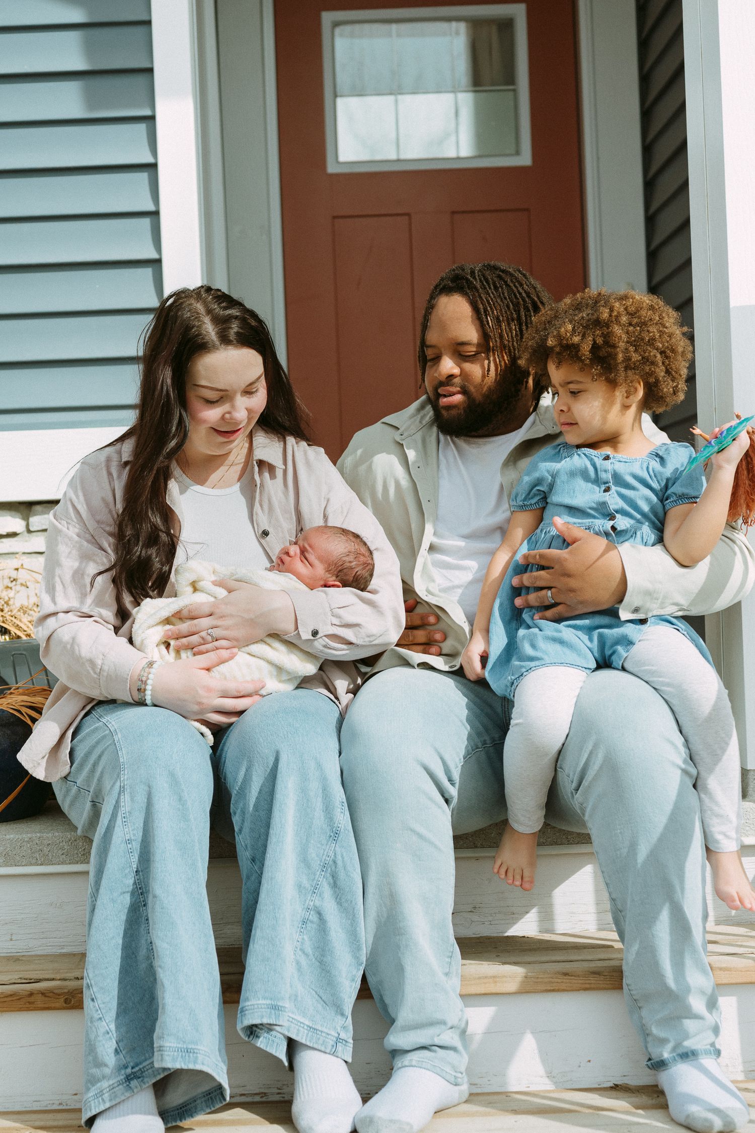 A happy interracial family sits on porch steps, parents holding a newborn and toddler in blue dress.