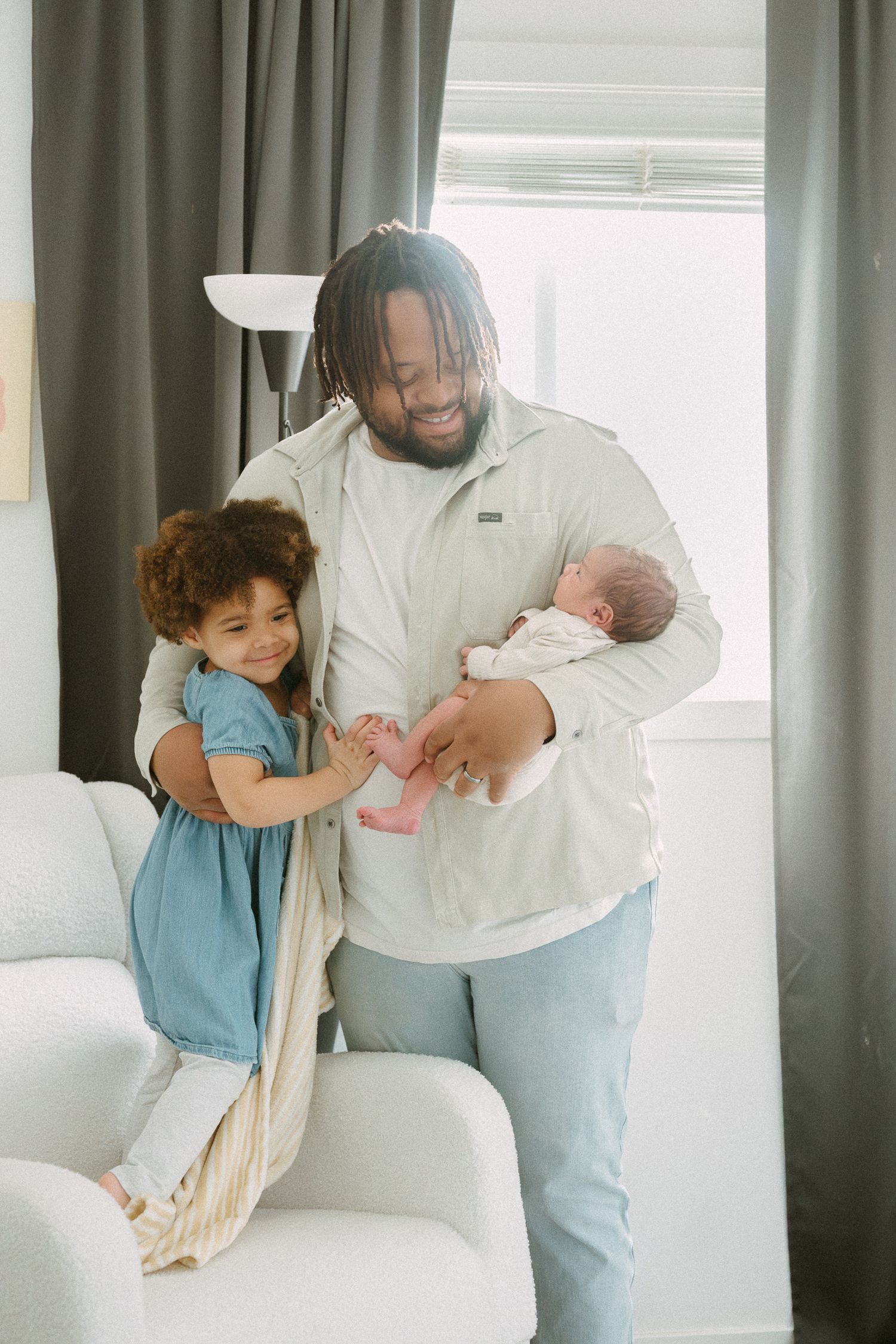 Smiling father holding newborn baby while young toddler girl in blue dress reaches for sibling in bright living room.