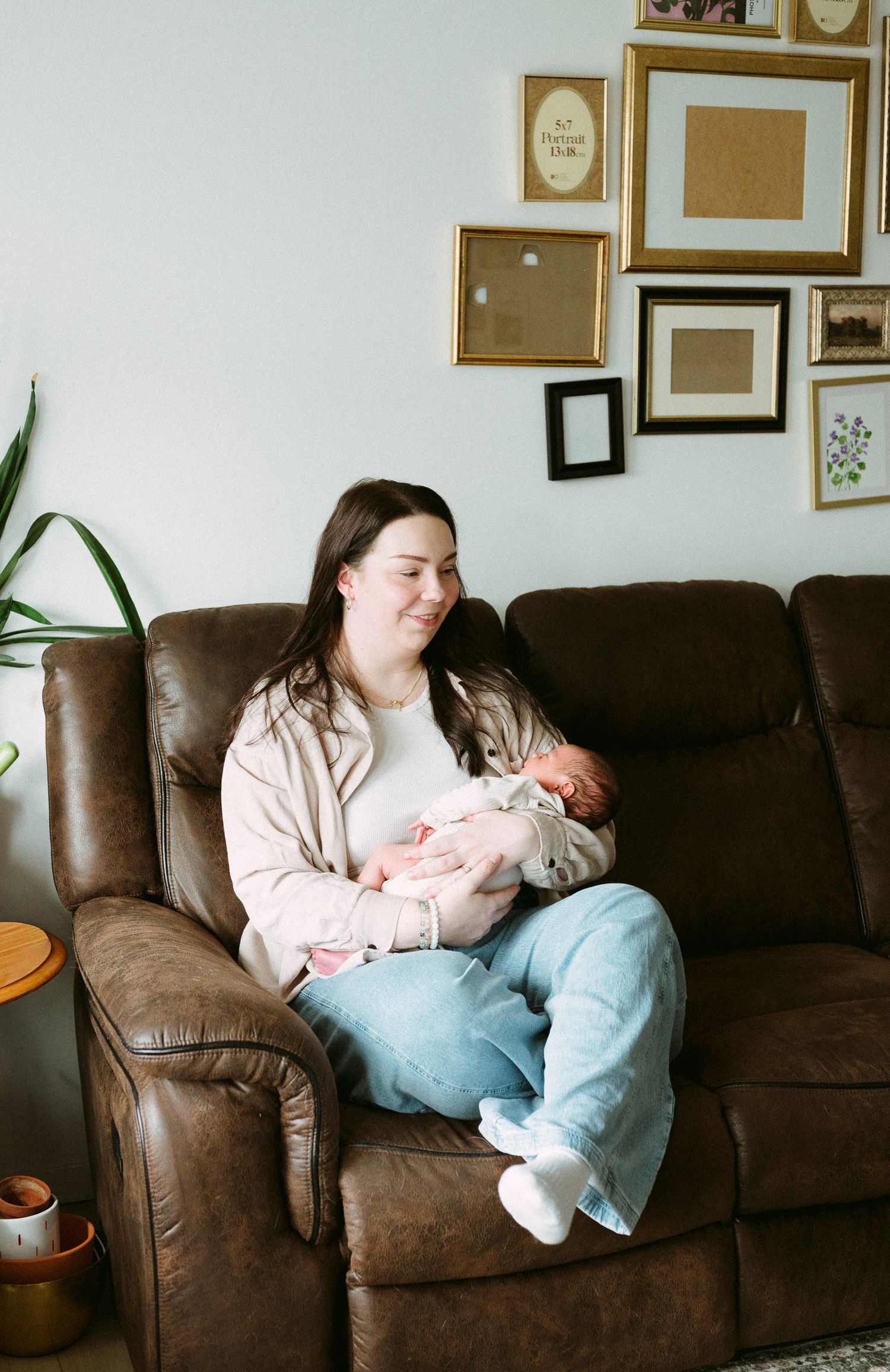 A young mother sits in a brown leather recliner, smiling down at her newborn baby in a cozy living room.