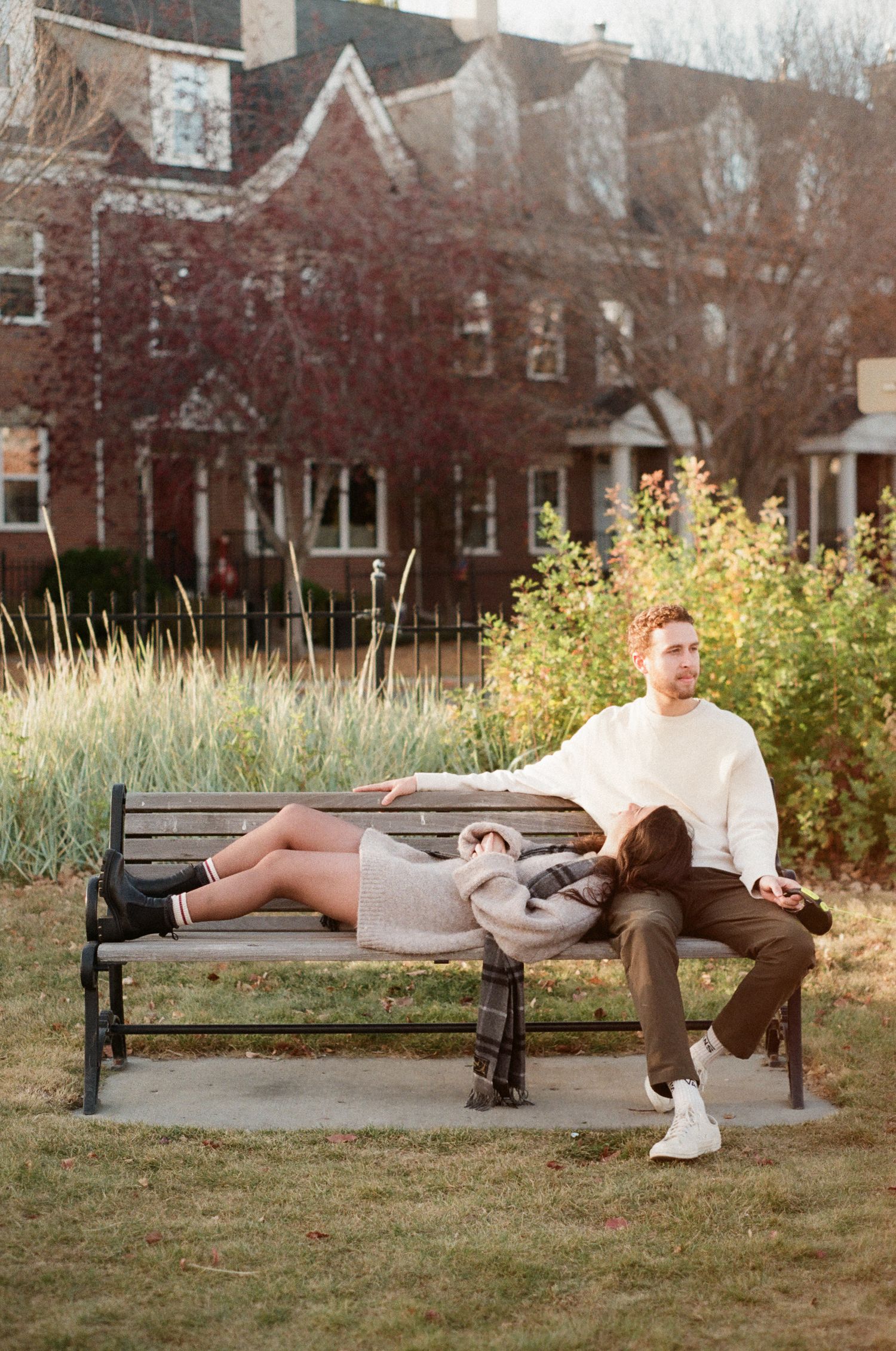 Couple relaxing on park benches during golden hour with brick buildings in background.