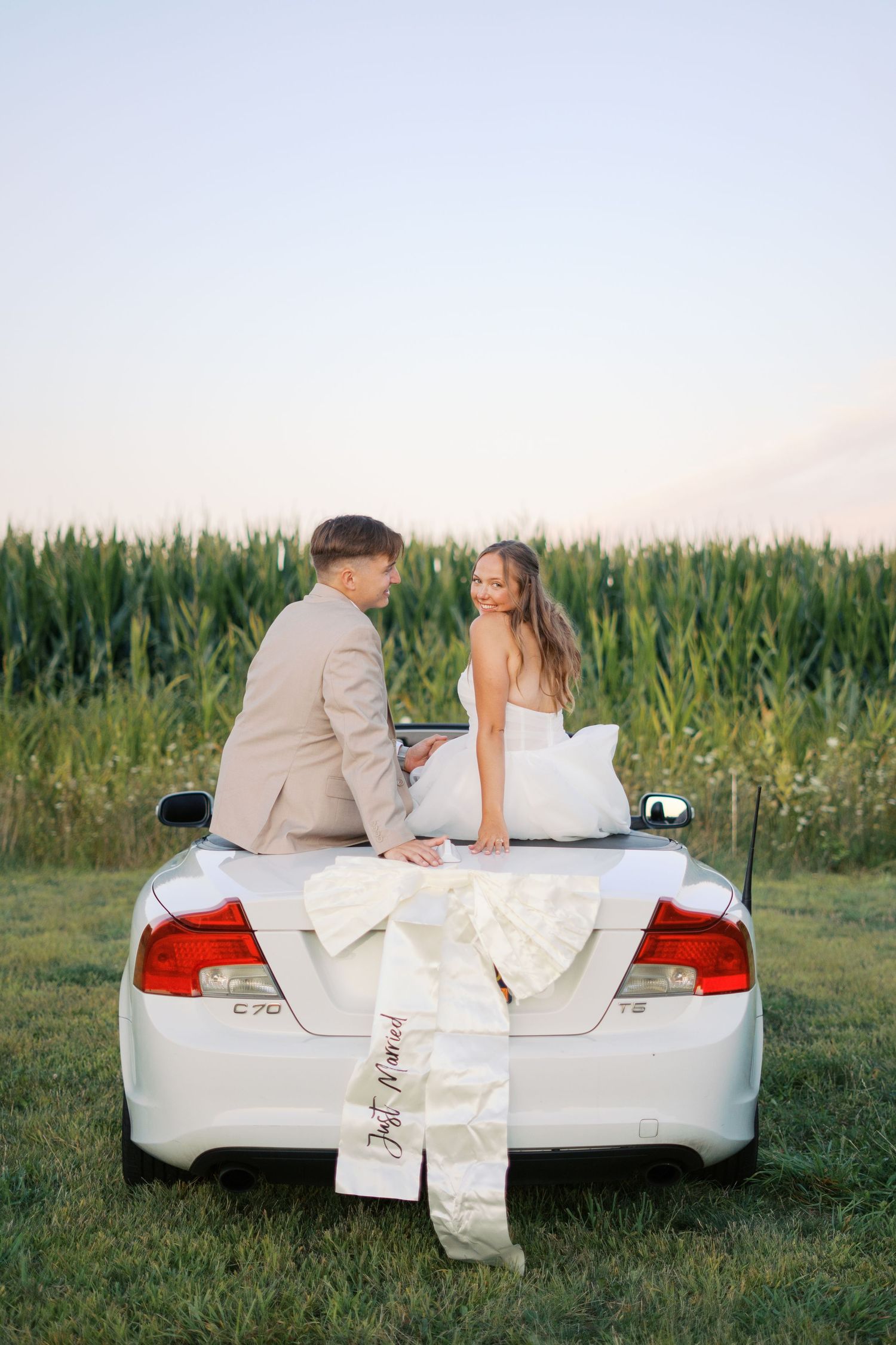 Wedding couple poses romantically on white car trunk near cornfield at sunset.
