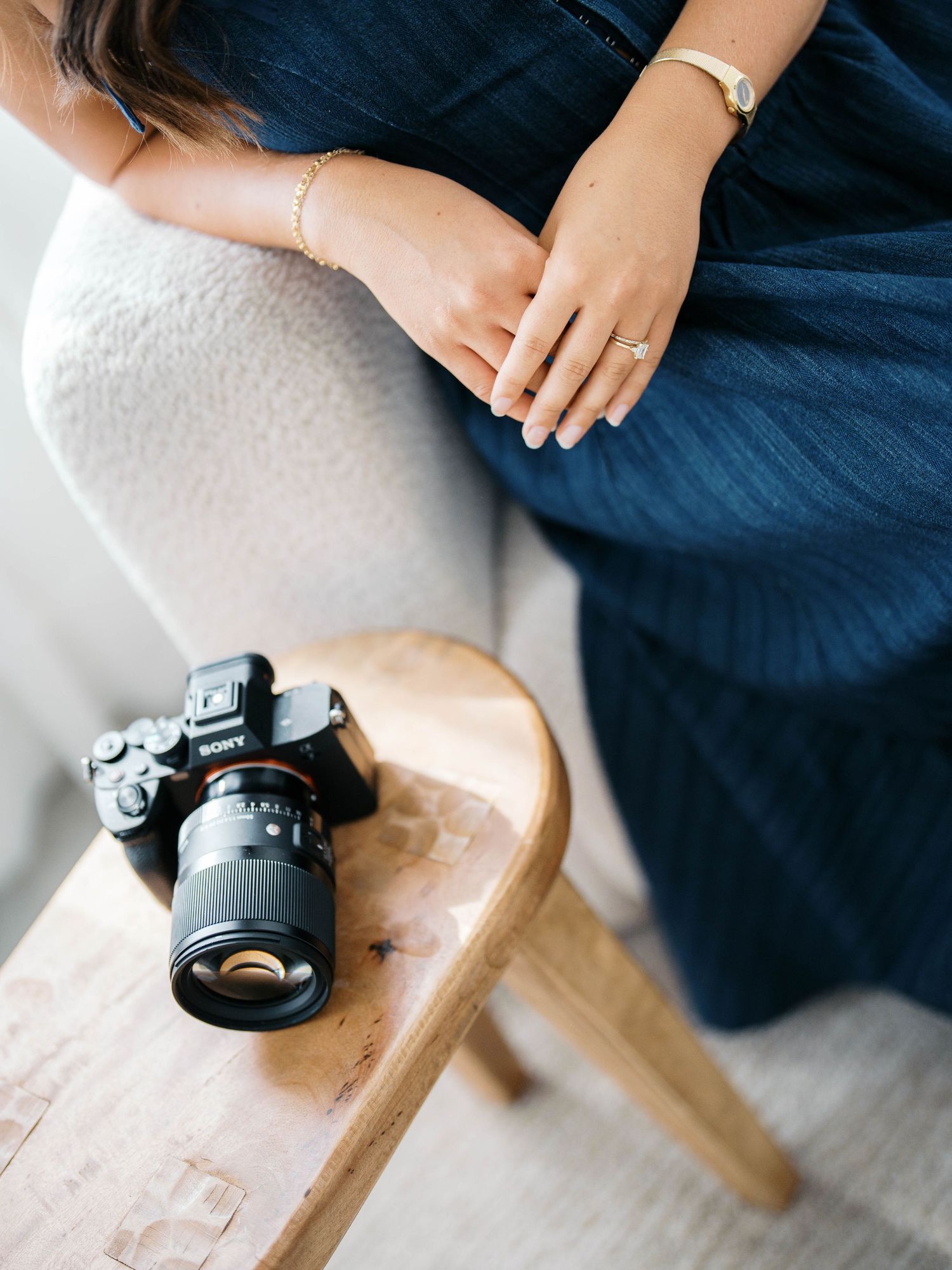 Woman in navy dress sitting beside a wooden stool with a Sony mirrorless camera, wearing gold jewelry.