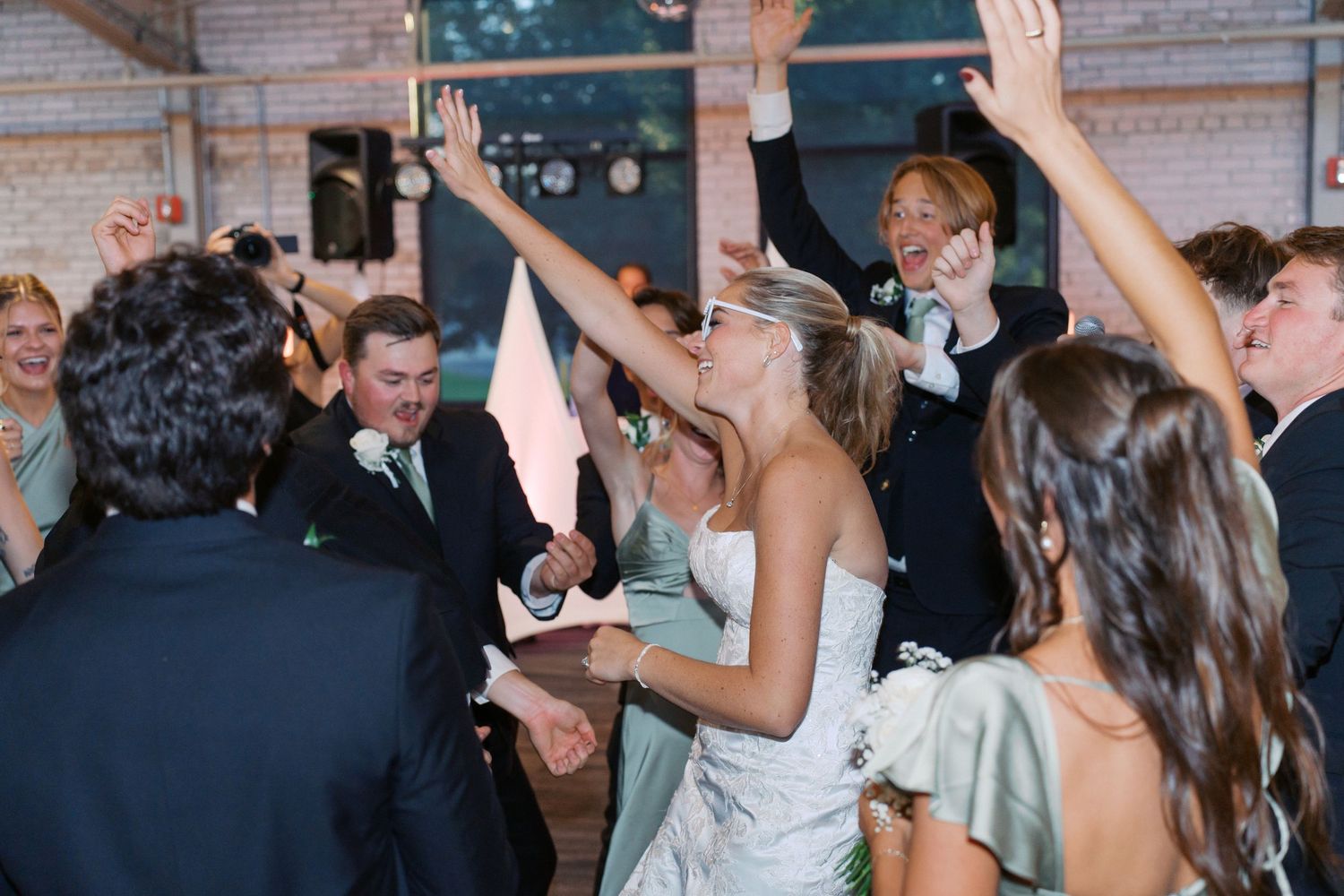 Wedding reception guests dance energetically under string lights in an industrial venue.