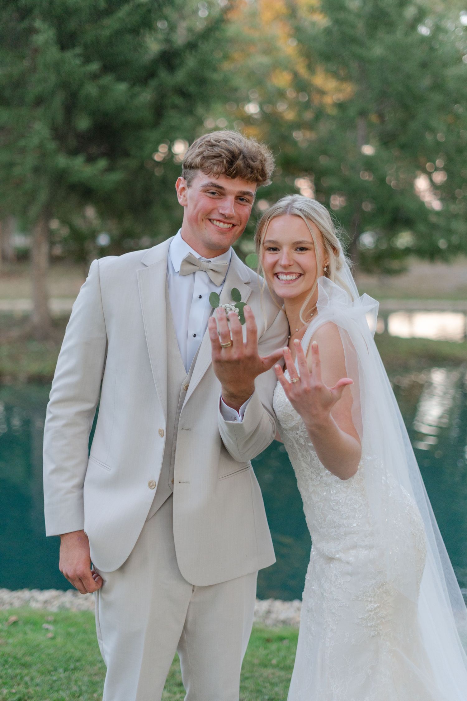 Newlyweds in cream suit and white dress show off their rings by a lake.