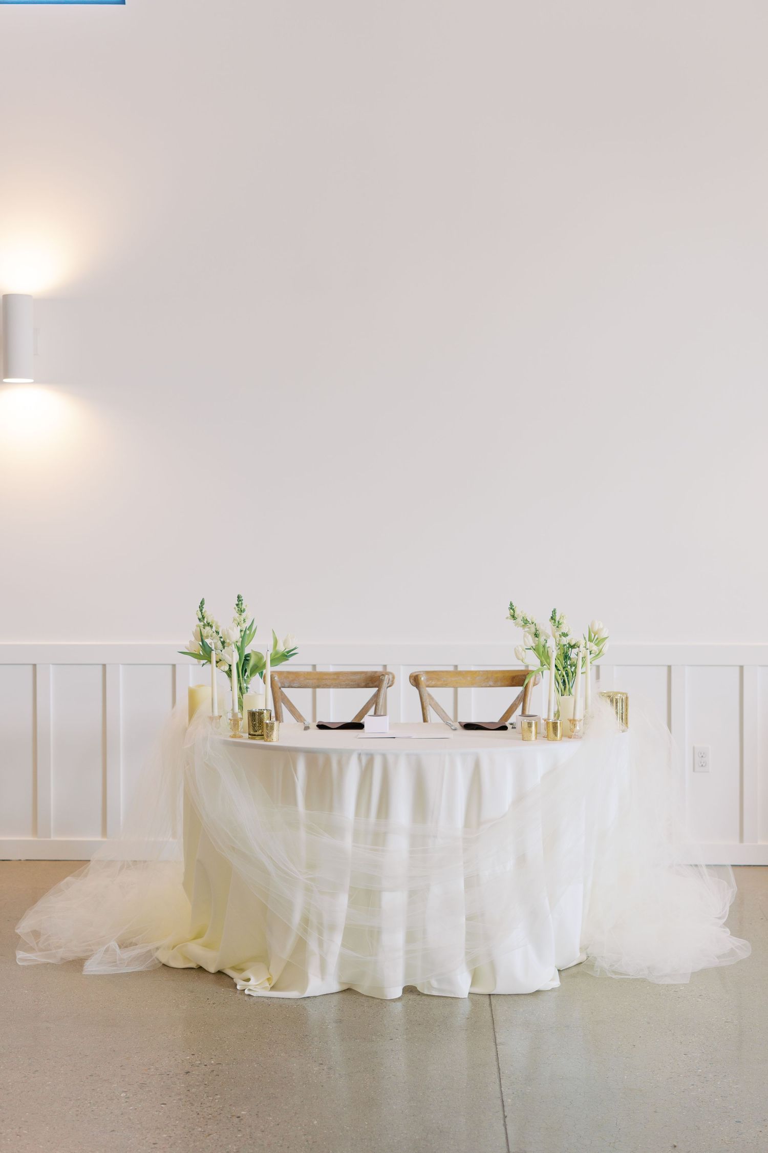 Elegant sweetheart table with white linen, gold accents, wooden chairs, and green floral arrangements in a bright white venue.
