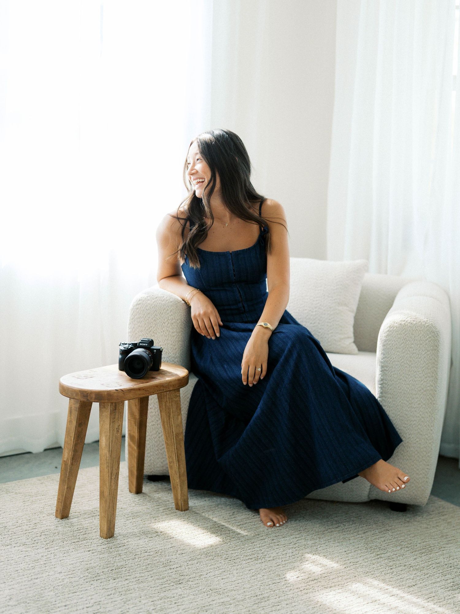Smiling woman in navy dress sitting on white armchair beside wooden stool with camera in bright airy room.