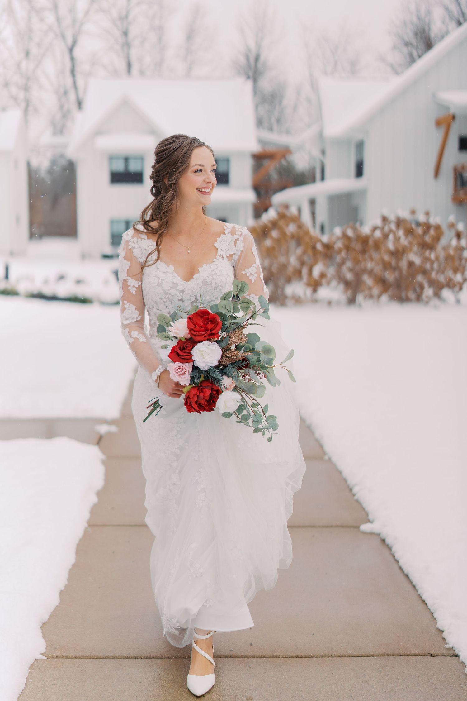 Bride in lace gown holds red and white bouquet while posing in snowy winter setting.