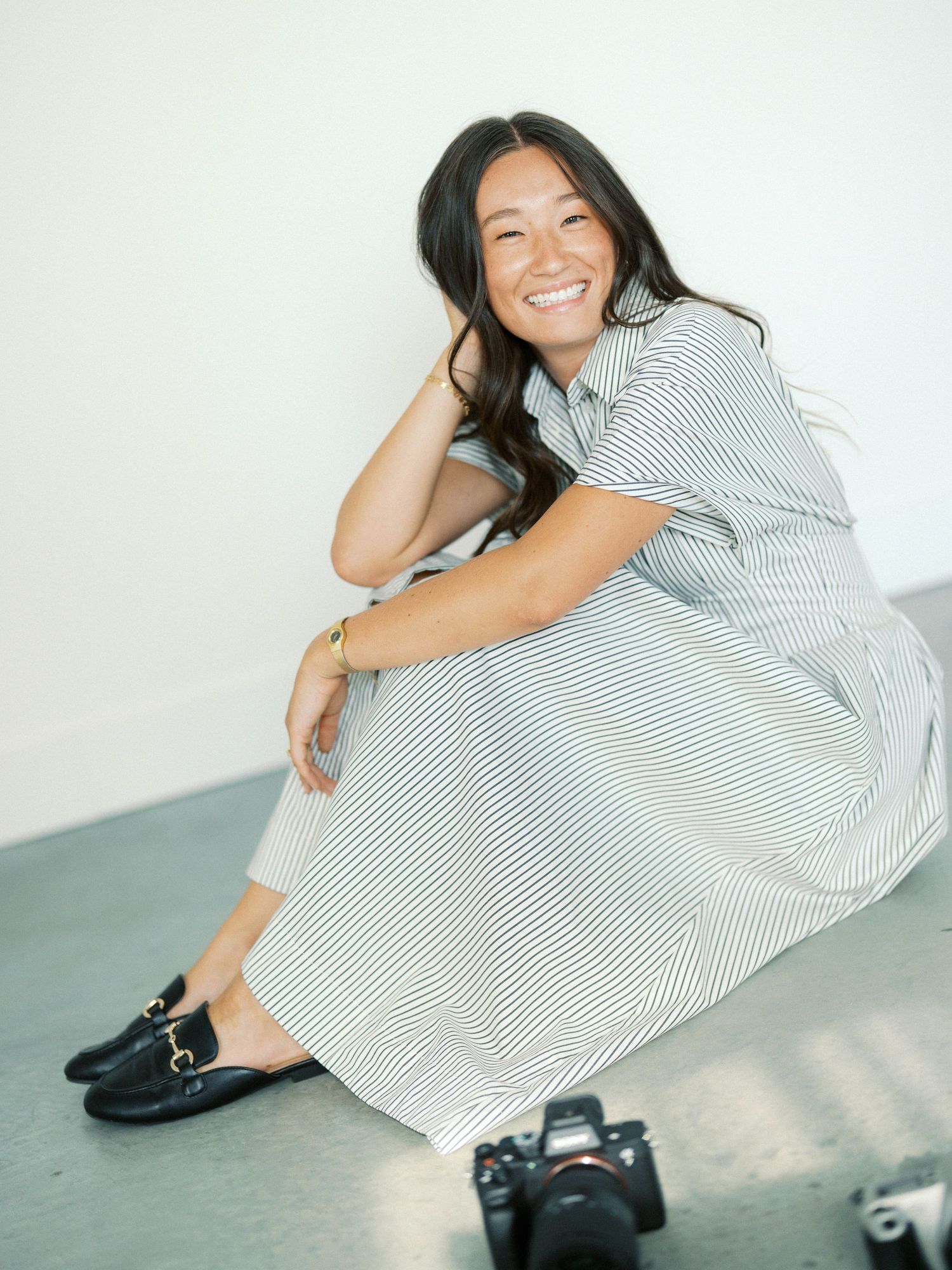 Smiling woman in striped dress and black loafers sits on floor near camera in bright studio.