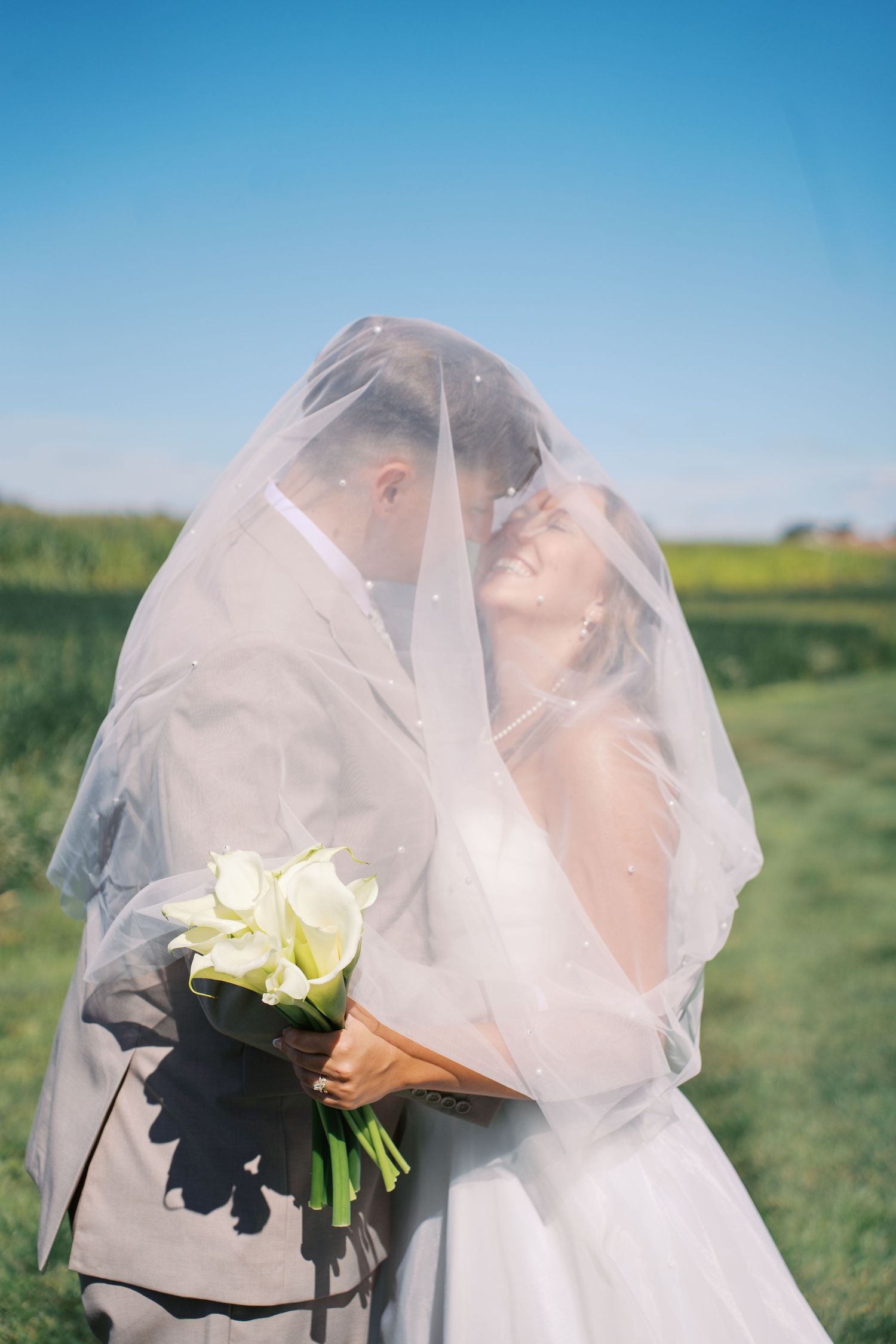 A bride holds a white bouquet under her flowing veil against a bright blue sky.