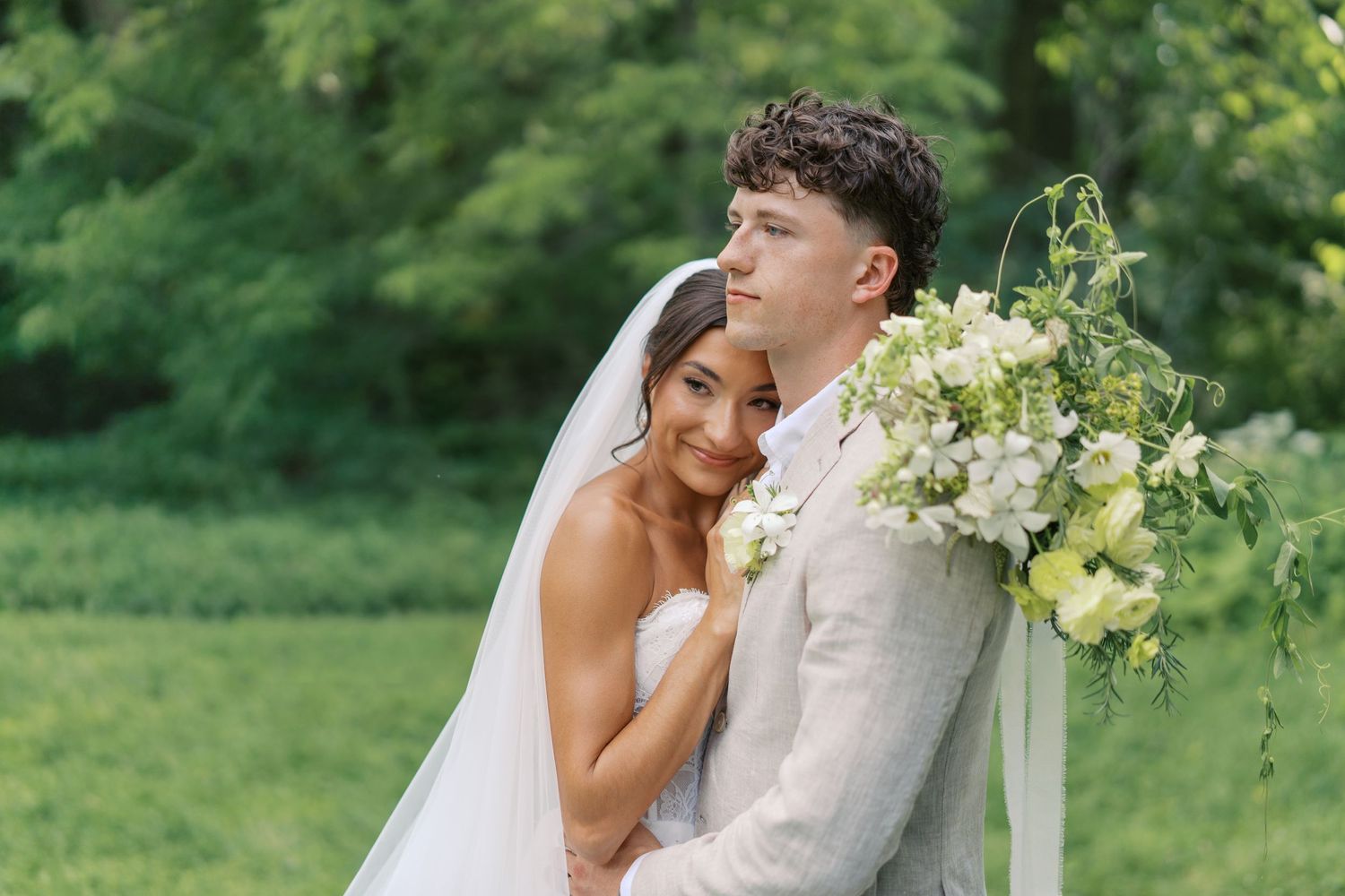 Bride in white gown and veil embraces groom holding lush floral bouquet outdoors at elegant garden wedding.