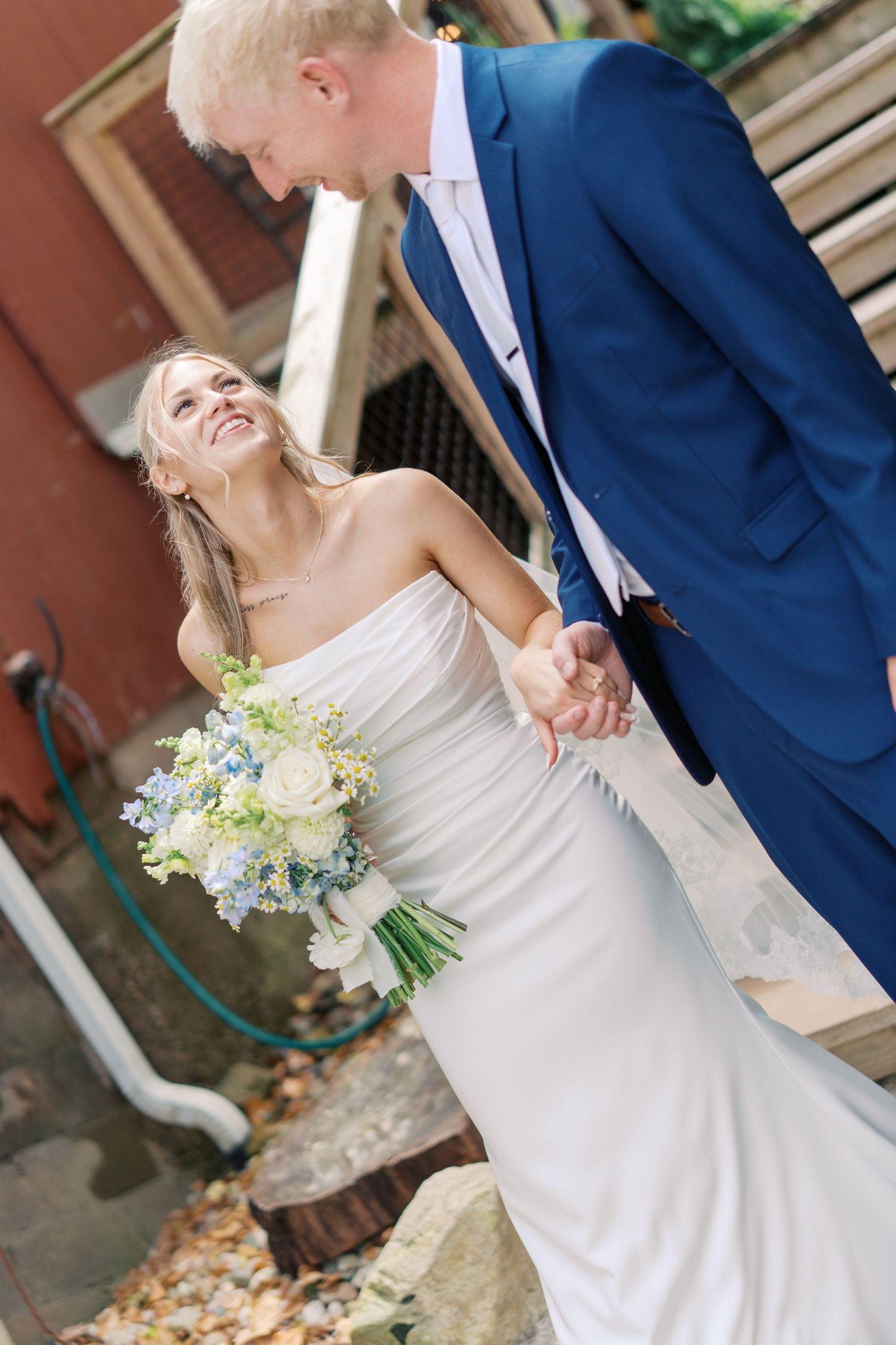 Bride in white strapless gown holds blue floral bouquet, smiling up at groom in navy suit while holding hands outdoors.
